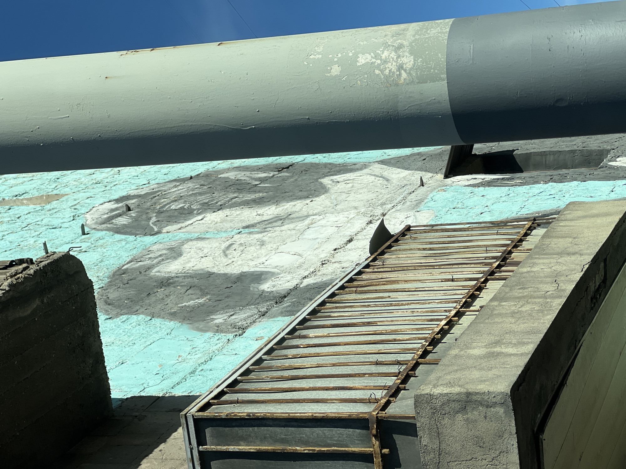 The image captures an exterior scene, potentially an industrial or infrastructural setting. A large, cylindrical pipe, painted in shades of gray and pale green, dominates the upper portion of the frame. Below the pipe is a concrete structure featuring a mural in faded shades of blue, white, and gray, possibly depicting abstract forms or a landscape. A weathered metal grate or walkway spans a gap in the concrete structure, suggesting a maintenance access point or a utility pathway.
Based on the location of Yerevan, Armenia, this scene is highly suggestive of old soviet buildings. More likely, an abandoned factory building. No people or other subjects.