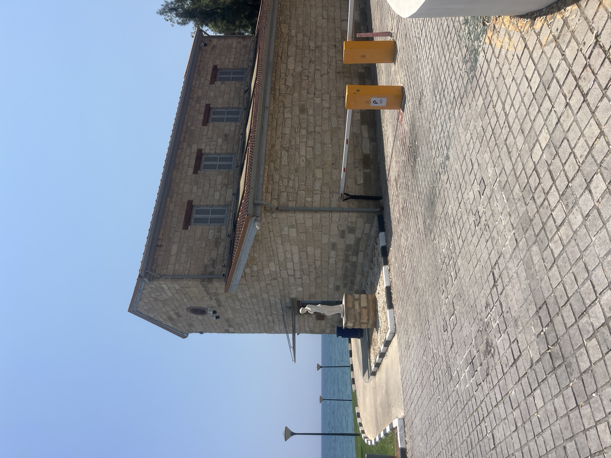 The image shows a bright, sunny day in Agios Tychona Community, Cyprus. The scene features a stone building with a red tiled roof, several shuttered windows, and a circular window on the side. In front of the building, a white statue of a person stands on a stone pedestal. To the left of the building, there are three lampposts along the coast, with the sea and a clear blue sky in the background. The ground in the foreground is paved with cobblestones. There are two yellow parking barriers with a "P" symbol and an arrow on them. The overall environment appears to be a public space near the sea.