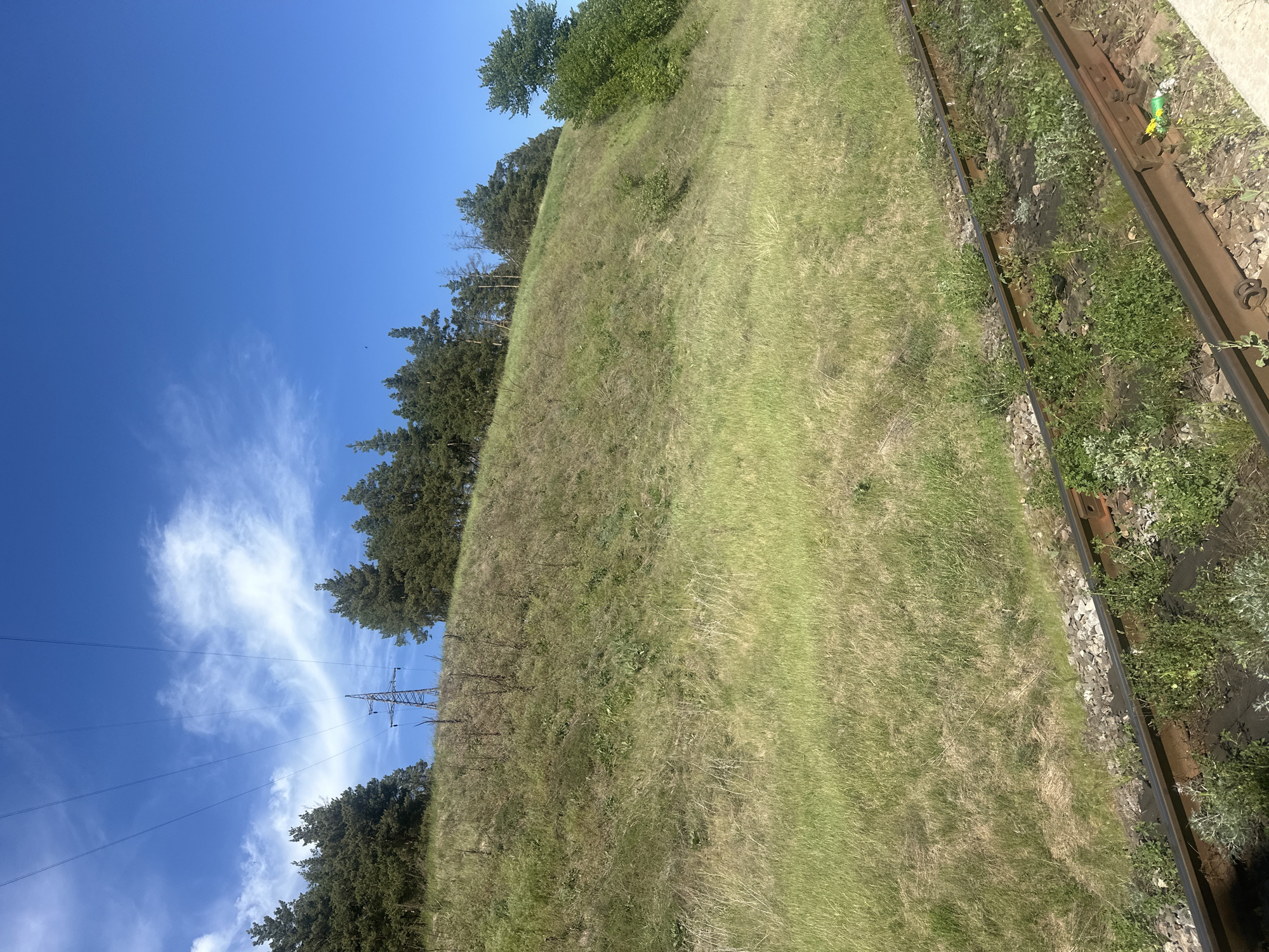 The image depicts a grassy hillside adjacent to a section of railway track. The sky is blue with scattered clouds and electrical power lines are visible in the background, near a line of trees along the hill's crest. The image location is in Pakhynia, Ukraine. The hillside is covered in green grass and some scrub vegetation. The railway track consists of two parallel metal rails, with ballast. There are no people visible in the frame.