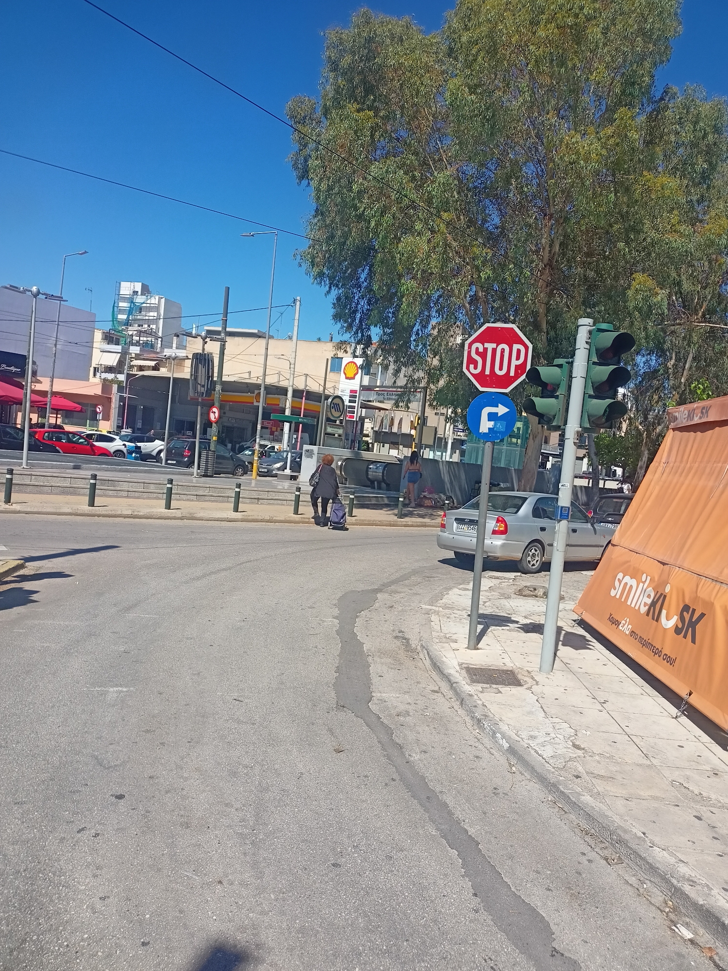 A medium shot captures an urban street scene under a clear blue sky, with a large tree to the right. In the foreground, a paved road transitions to a sidewalk. On the sidewalk, a person with dark hair and dark clothing is pulling a blue wheeled cart. To the right, a traffic light pole displays a red octagon "STOP" sign above a circular blue sign with a white arrow indicating a right turn. Below these, the traffic lights themselves are visible. Further right, an orange awning displays the word "smile kiosk" in white text, with Greek lettering below. In the background, several buildings are visible, including a Shell gas station with a prominent yellow and red logo, and other structures suggesting a commercial area. Cars are parked and driving on the road in the mid-ground.