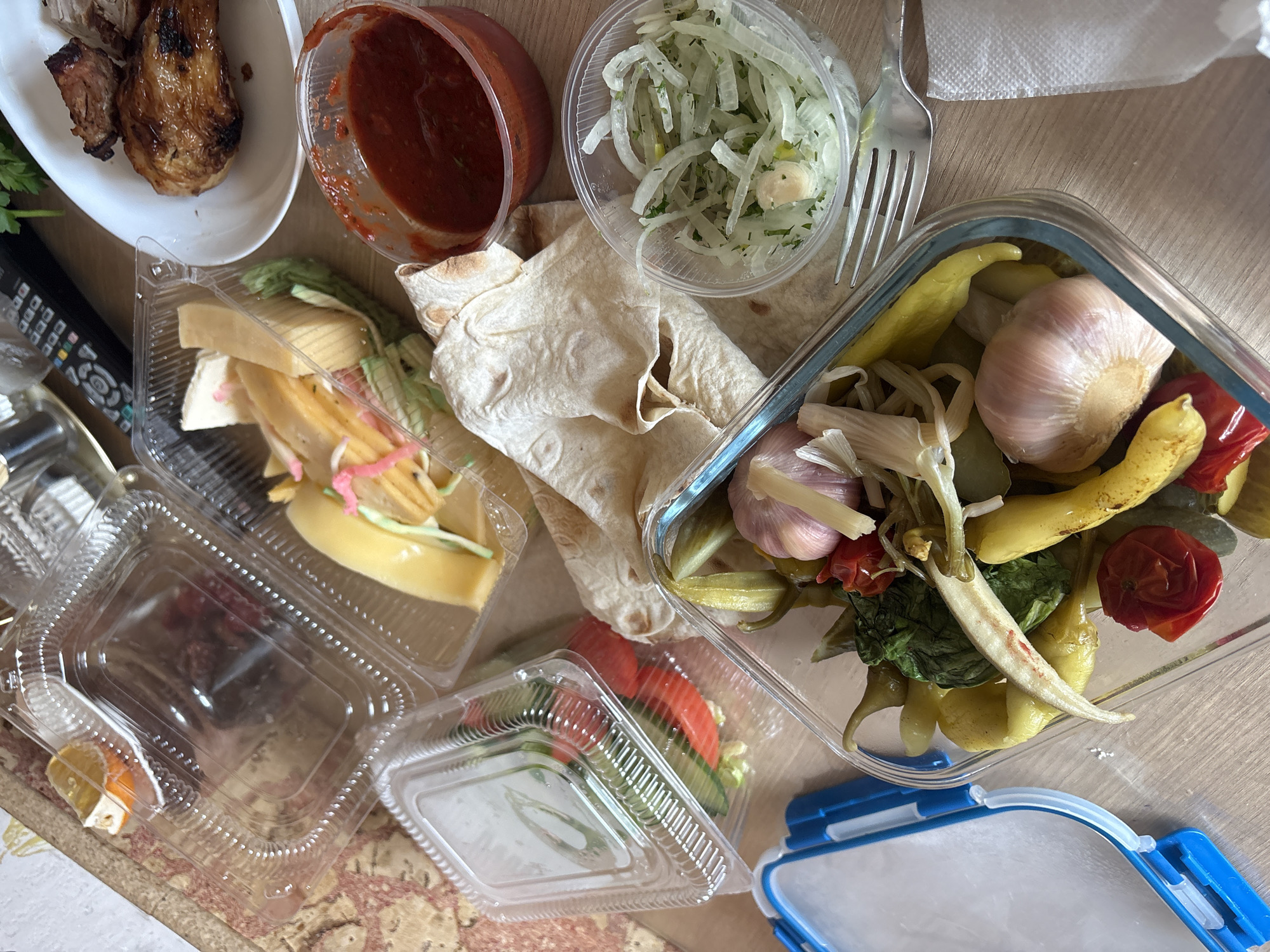 The image displays a spread of food items in various containers on a light brown, patterned table, indicative of a casual meal, likely takeout or a relaxed gathering in Krasnoyarsk, Russia.
The primary subjects are diverse food items and their packaging. In the upper left, a white oval dish holds pieces of grilled meat, resembling shashlik or grilled chicken, with visible char marks. Next to it, a clear plastic cup contains a red sauce, possibly ketchup or ajika. Another clear plastic cup holds thinly sliced pickled onions with herbs. A piece of flatbread, likely lavash, is placed in the center.
Below the grilled meat, a clear plastic container holds slices of cheese, some white and some yellow, with some shredded vegetables and pink-colored garnish. To its right, another clear plastic container appears to hold a simple salad with sliced tomatoes and cucumbers.
Dominating the right side is a glass container with a blue-rimmed base, filled with an assortment of pickled vegetables, including whole garlic cloves, yellow peppers, small red pickled tomatoes, and possibly other green pickled items like okra or green beans. A silver metal fork and a white paper napkin are also visible on the table.
Other objects include a black remote control partially visible on the left and the base of a clear glass or bottle. The food is arranged somewhat haphazardly, suggesting a casual, post-delivery setup. No specific brand names or detailed text are clearly legible on any of the containers or the remote control. The overall scene suggests a hearty, informal meal.