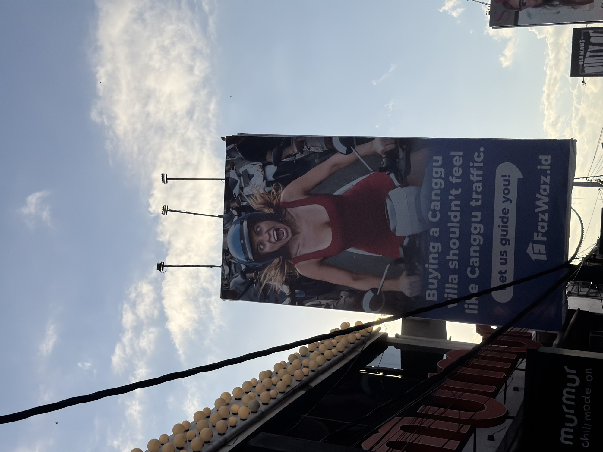 The image is captured from below, looking upwards towards a billboard advertising FazWaz.id, featuring an image of a woman wearing a helmet on what appears to be a motorcycle or scooter. The billboard copy reads, "Buying a Canggu Villa shouldn't feel like Canggu traffic. Let us guide you! FazWaz.id."
Below the billboard is a roofline with a string of globe lights. The branding "murmur chill mode: on" is partially visible, indicating the name of a business beneath the billboard. The background shows a sky with clouds.
Another sign with the words "OLD MAMS" on it is partially visible in the top right corner of the frame. The location of this image is North Kuta, Indonesia.