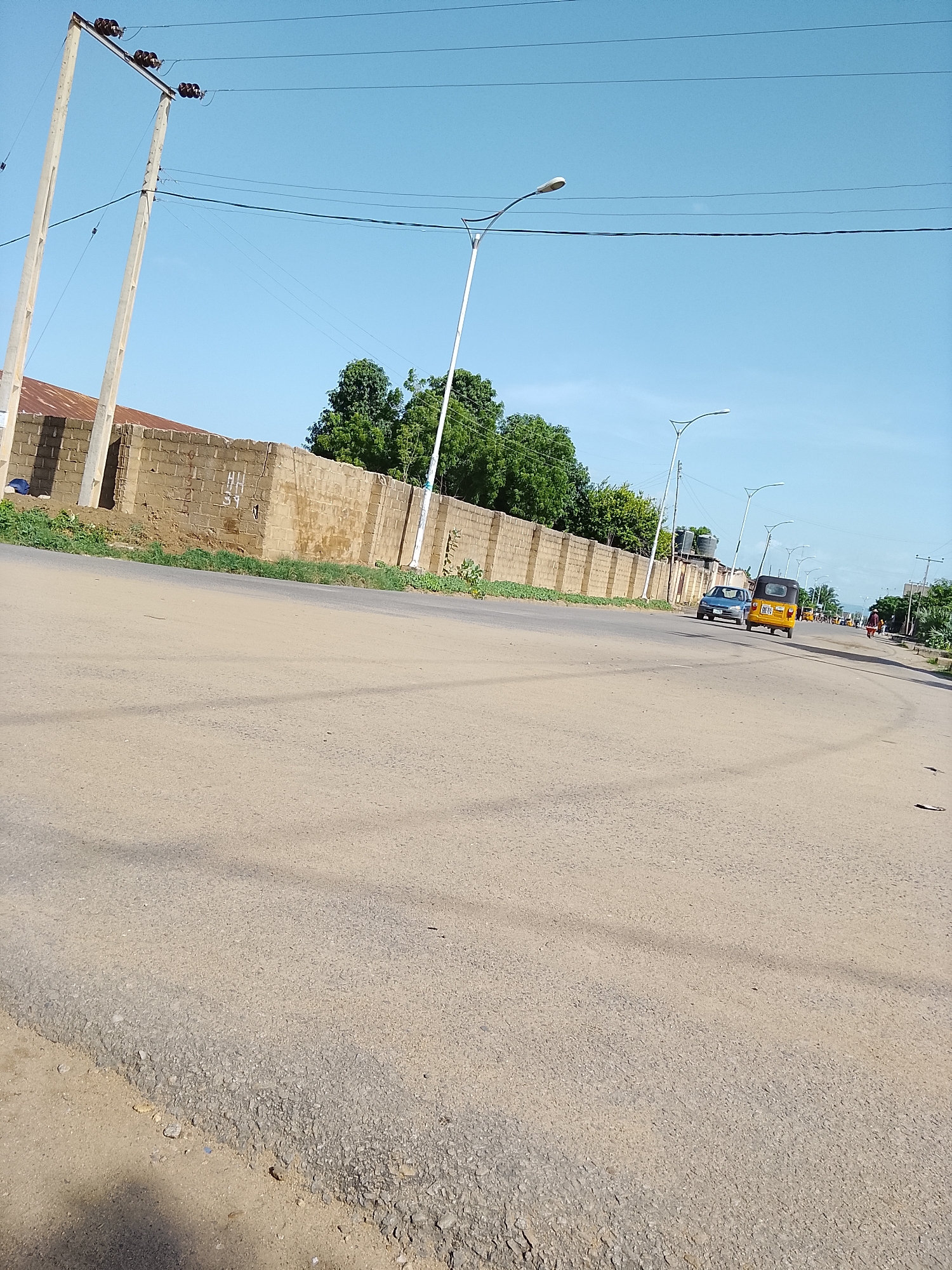 The image depicts a street scene in Wauro Leggal, Nigeria, on a clear day under a blue sky. In the foreground, a paved road shows signs of wear and tear, with some areas of exposed aggregate. A shadow of a person or object is cast across the road. On the left side of the road, a brick wall with a corrugated metal roof is visible, adjacent to a grassy verge and two tall concrete utility poles with electrical wires.
Further down the road, streetlights line the asphalt. A yellow autorickshaw with the license plate "BGT 95" is traveling in the same direction as the viewer, followed by a dark-colored car. Several other vehicles and a person with a cart are visible in the distance, suggesting moderate traffic. Trees and more utility poles are scattered along the right side of the road. The overall environment suggests an urban or semi-urban setting.