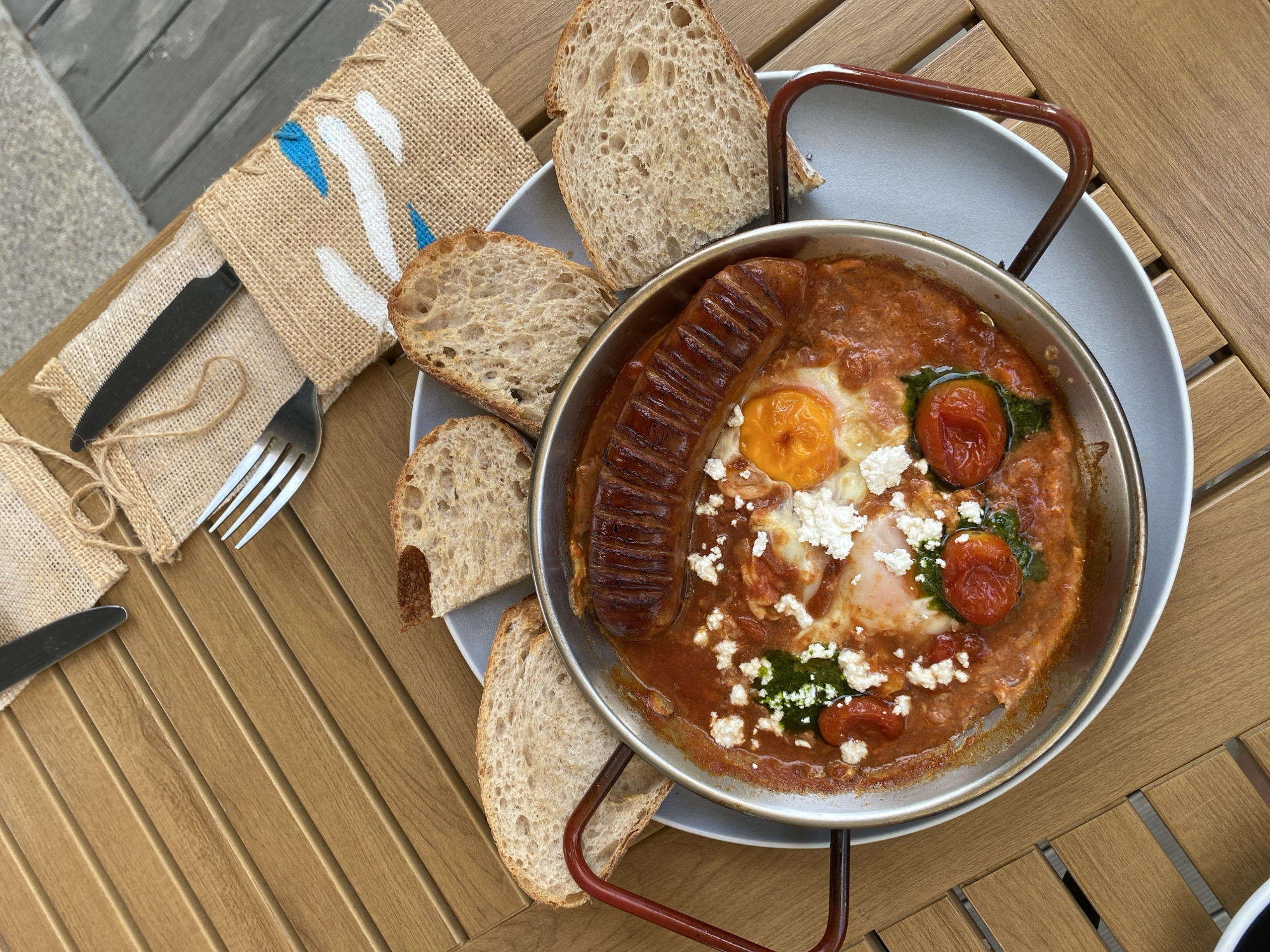 A high-angle shot captures a culinary composition featuring shakshuka served in a metal pan with a handle, situated on a grey plate. The plate rests on a wooden-slatted table. The dish includes a sausage, eggs, tomato sauce, feta cheese, and basil pesto. Four slices of sourdough bread are arranged around the pan. A burlap napkin with white and blue markings, a knife, and a fork are to the left of the plate. The location is Choeng Thale, Thailand.