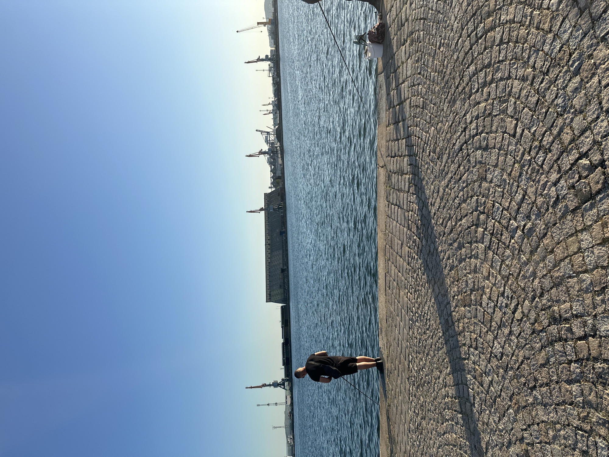 The image displays an outdoor scene at a port or harbor on a clear, sunny day. A vast expanse of deep blue water, showing ripples on its surface, dominates the left side of the frame. To the right, a cobblestone quay or promenade runs alongside the water. The grey cobblestones are arranged in an arched pattern, with long shadows stretching across them from the right, suggesting morning or late afternoon sun.
Two individuals are present. In the mid-ground, a person wearing a black t-shirt and black shorts stands near the water's edge, leaning forward as if observing or attending to something in the water. Further to the right, partially visible, another person is seated or crouching on the cobblestones next to a white bucket. A fishing rod extends from their position towards the water, indicating they are fishing.
In the background, across the water, an industrial landscape is visible. Numerous large cranes of various types line the distant shore, alongside dark, rectangular structures that appear to be port buildings or parts of large vessels. The clear blue sky is largely cloudless. There is no visible text in the image.