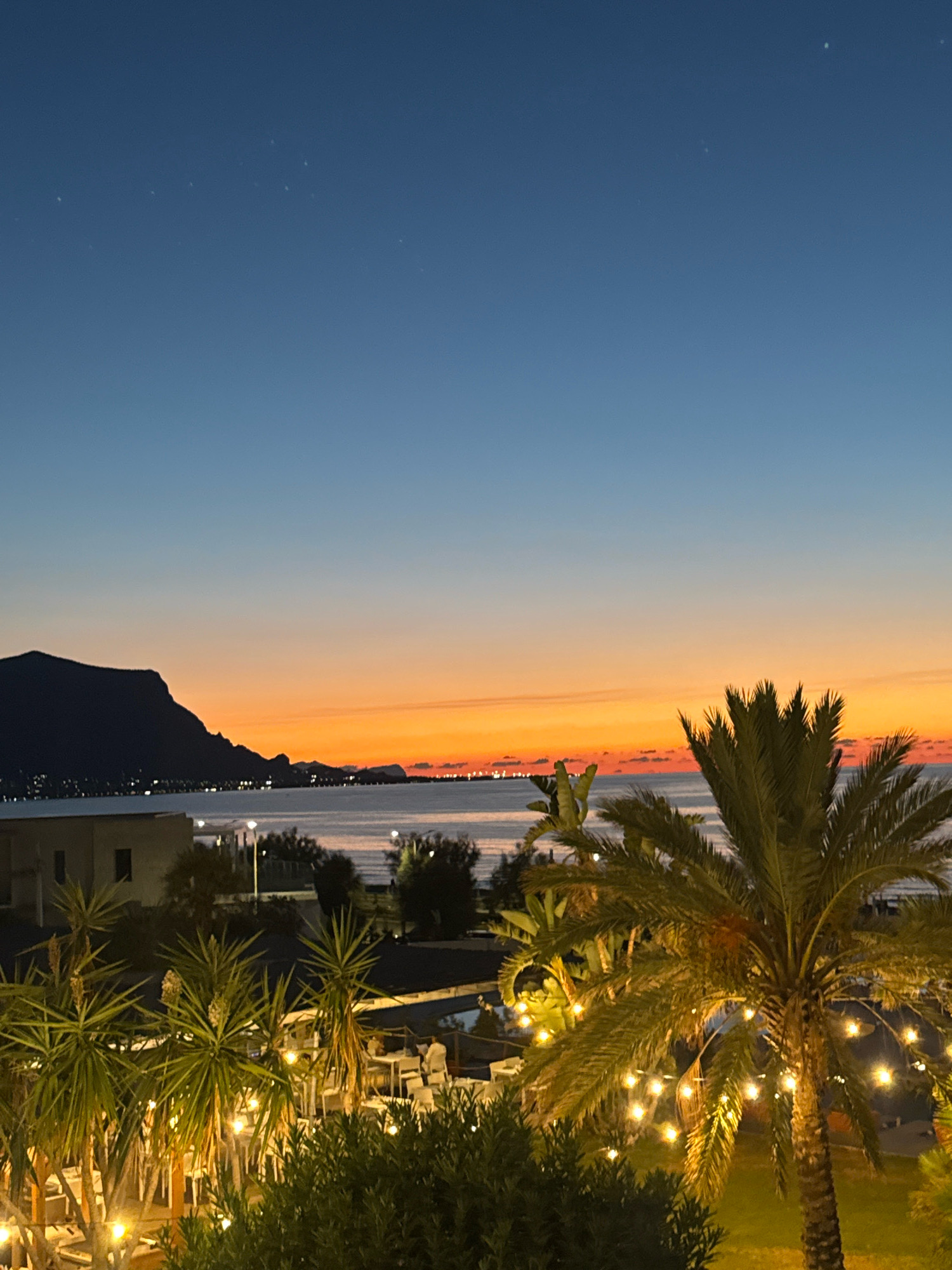 This is a nighttime photograph taken from a balcony or elevated position overlooking the sea at dusk. The sky transitions from a deep blue at the top to a vibrant orange and pink horizon, indicating the sun has recently set. In the distance, silhouetted against the colorful sky, is the mountainous coastline of Isola delle Femmine, Italy. The water is calm and reflects the distant lights from the town.
In the foreground, several palm trees and other lush green foliage are visible. One large palm tree in the right foreground is adorned with a string of warm-toned fairy lights, which are illuminated. Behind the palm trees, there appear to be outdoor seating areas with tables and chairs, suggesting a restaurant or dining establishment. Some of these areas also have lighting. A few indistinct figures can be seen in the background, possibly patrons or staff of the establishment. The overall atmosphere is serene and inviting, capturing the twilight ambiance of a coastal location.