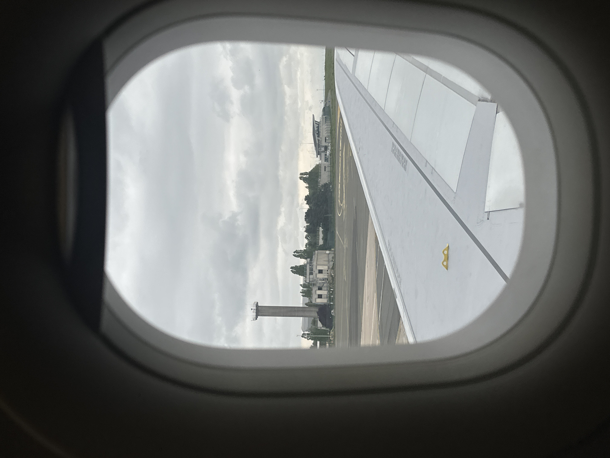 View through an airplane window showing the wing of the aircraft and the ground below, including a control tower. The location is Mauregard, France. The sky is overcast.