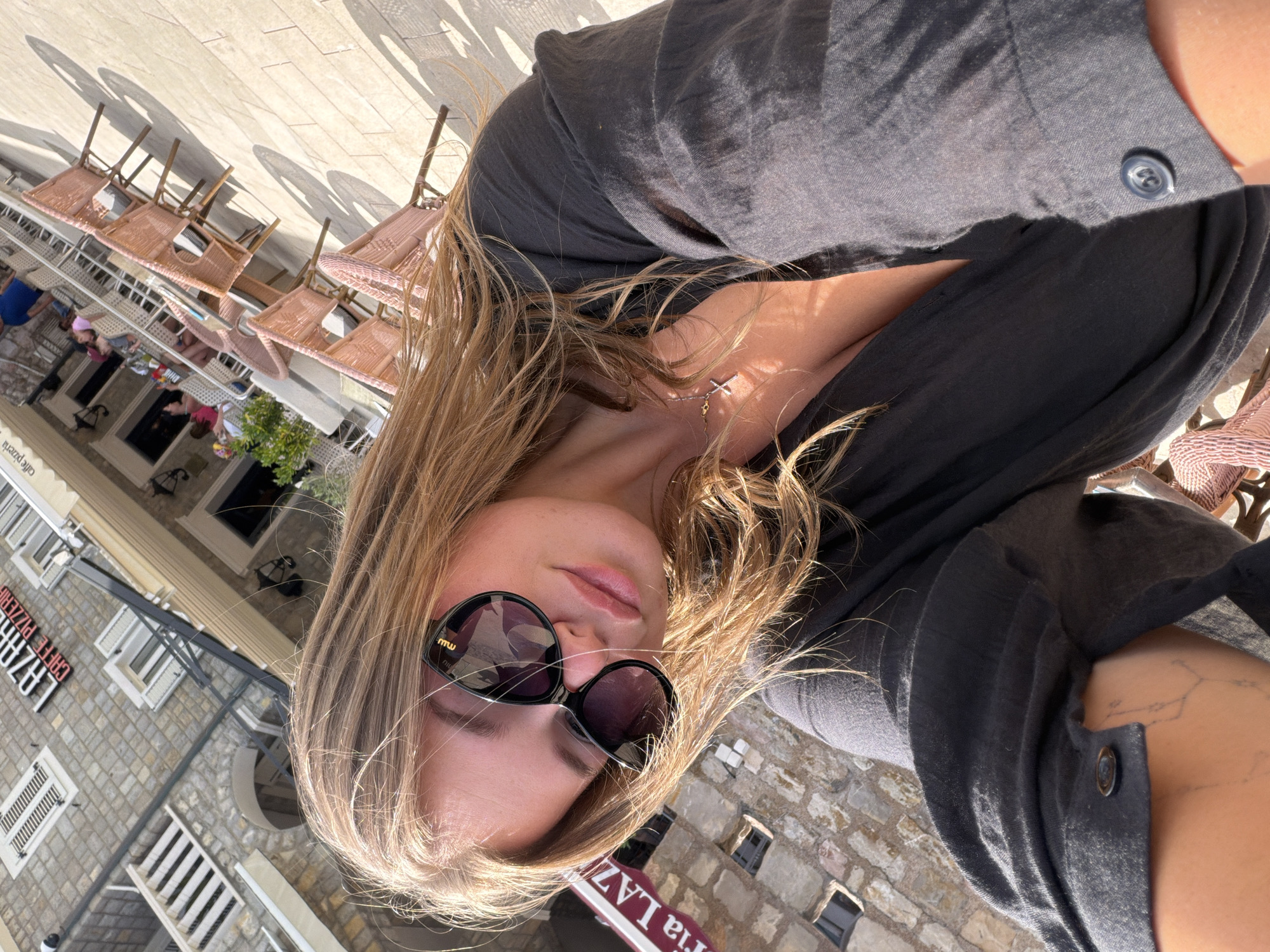 A woman is taking a selfie outdoors, with a cafe and building in the background. She has long, blonde hair and is wearing sunglasses and a dark, open-necked shirt. A small cross necklace is visible. The cafe appears to be located in Petrovac centar, Montenegro, with its name, "LAZAR", visible on a sign. The scene suggests a sunny day with shadows cast by chairs and umbrellas.