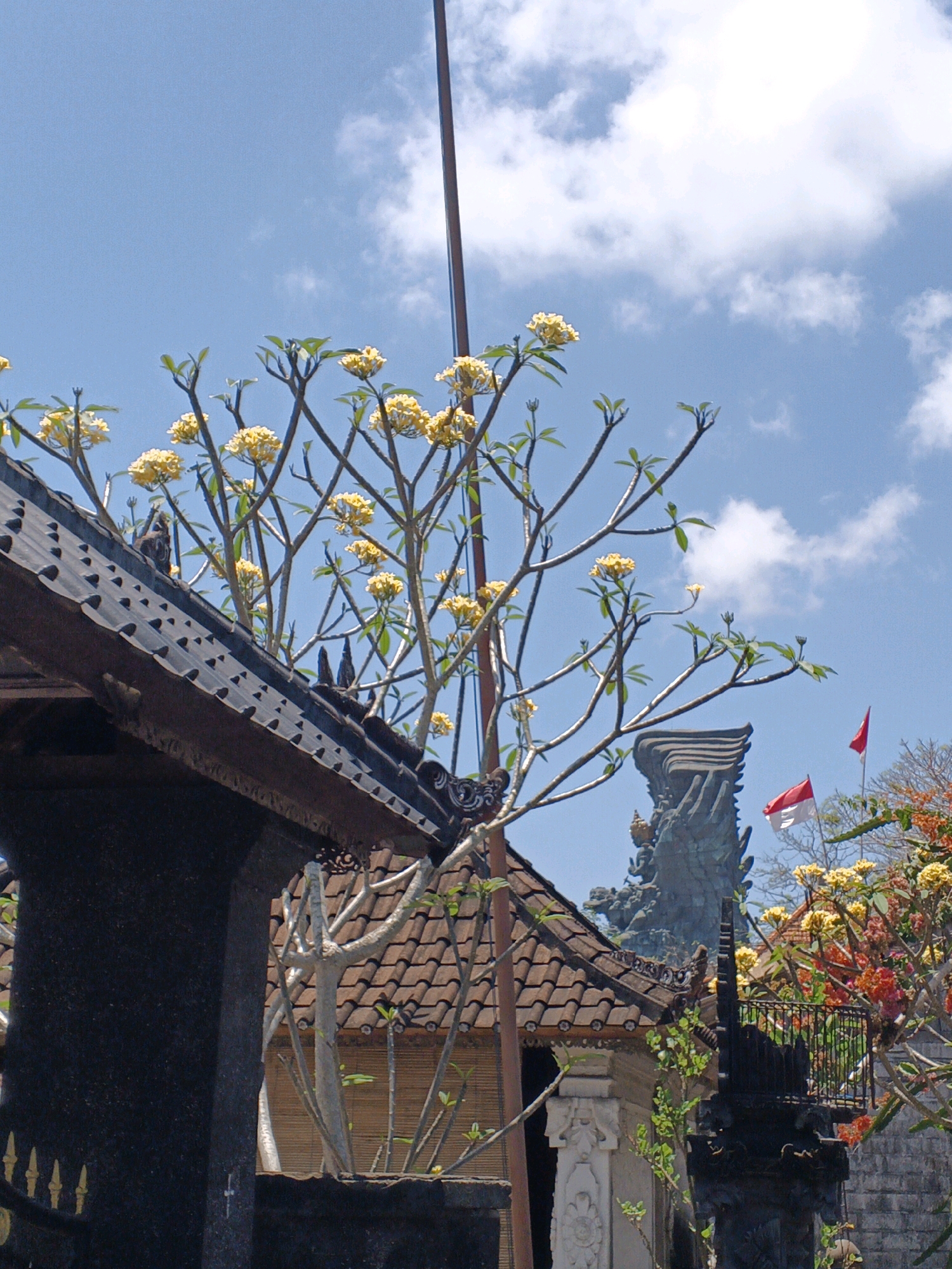 In Kuta Selatan, Indonesia, a balinese temple is viewed under a bright blue sky with scattered clouds. A tall, slender pole extends vertically through the frame. In the foreground, a dark tiled roof of a building juts into the lower left corner, adorned with decorative carvings. Above the roof, several branches of a tree with clusters of small yellow flowers are prominent. In the background, a large, dark statue of a mythical creature or deity is visible. To the right of the statue, the Indonesian flag, a red and white horizontal bicolor, is flown. Further to the right, more flowering trees with red and yellow blossoms can be seen, along with a stone wall and a black decorative metal railing. The overall scene depicts a cultural site bathed in sunlight.