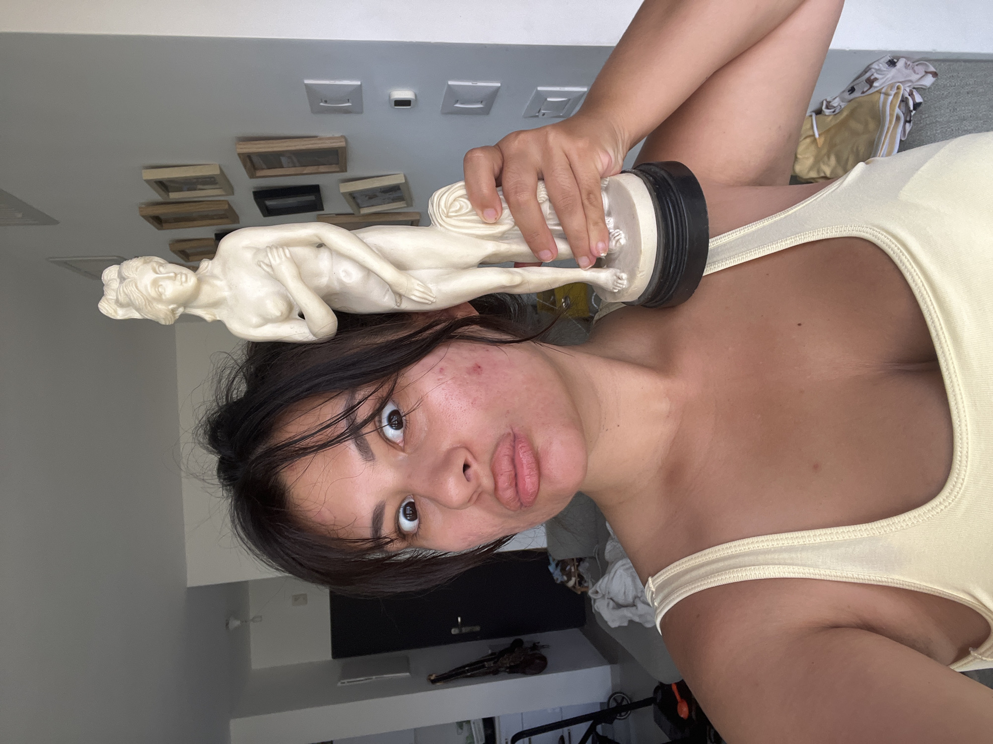 A woman with dark, messy hair and an exaggerated pout poses for a selfie in what appears to be a living room in Kiryat Bialik, Israel. She is holding a small, white statue of a nude woman with one hand. The statue depicts the woman with her hand covering her chest and a draped cloth around her legs. The woman in the foreground has fair skin with visible redness and blemishes on her cheeks and forehead, and her eyes are wide open and looking towards the camera. She is wearing a light yellow tank top. In the background, there is a wall with several framed pictures hanging on it, and a light-colored ceiling with a light fixture. A stroller can be seen on the left side of the image, and a couch with some folded clothes on it is visible in the lower right portion. The overall lighting suggests it is daytime.
