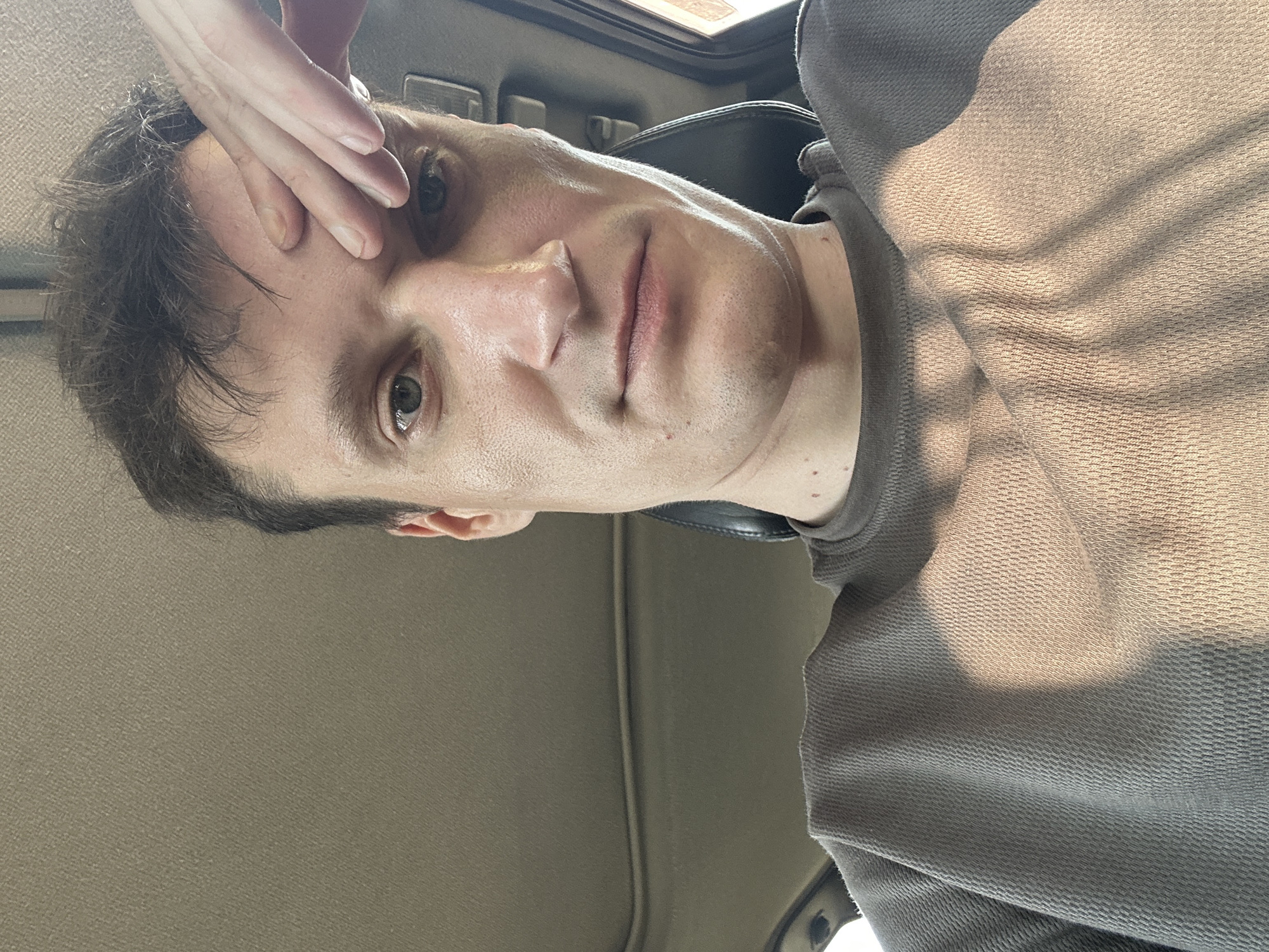 The image shows a close-up, selfie-style photograph of a man from the chest up, seated inside a vehicle, likely a car. He has dark hair and light skin, and his right hand is placed on his forehead, possibly indicating distress or contemplation. The man is wearing a dark grey, long-sleeved shirt with a textured fabric. Sunlight is casting shadows across his shirt and face, suggesting it is daytime. The interior of the car is visible, with parts of the ceiling and a headrest. There are no other people or objects in the frame that provide specific details about the location within Makiv, Ukraine, or any specific activities occurring. The overall impression is a candid, personal moment captured by the individual himself.