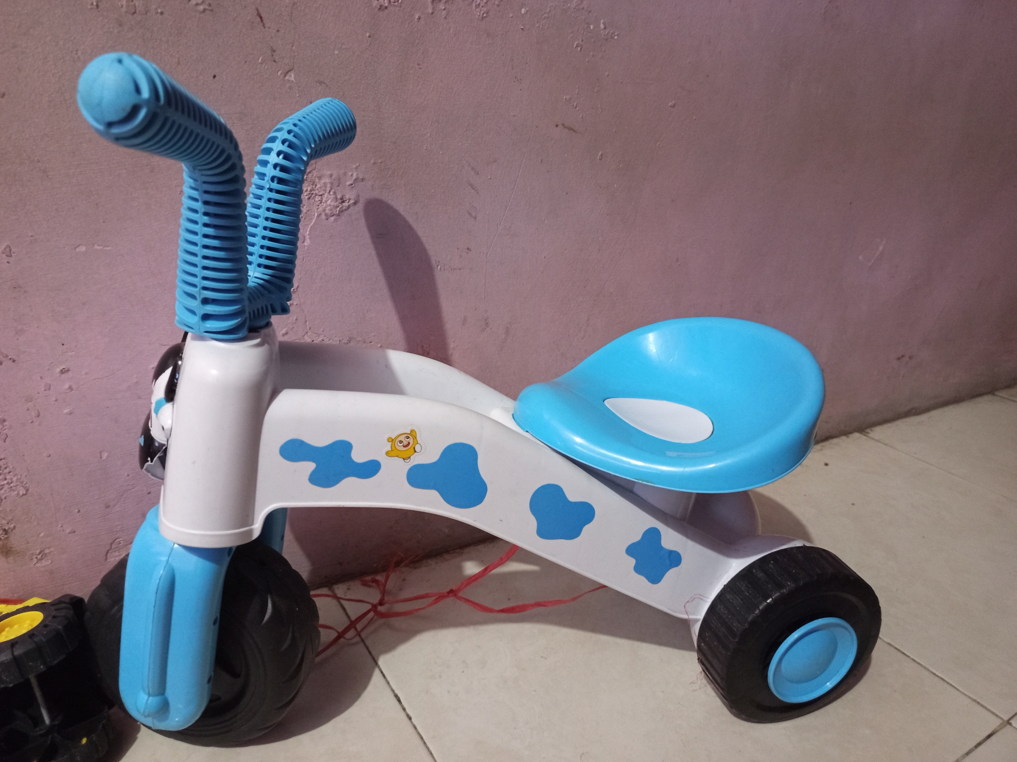 A blue and white plastic child's tricycle with cow-print decals and a yellow cartoon monkey sticker is positioned on a tiled floor against a pale pink textured wall. The tricycle has two blue handlebars, a white body, a blue seat, and three black tires with blue hubcaps. A red string is tangled underneath the tricycle's frame. To the left, the front wheel of another toy, likely a yellow and black wheel with a yellow rim, is visible. The lighting suggests an indoor environment.