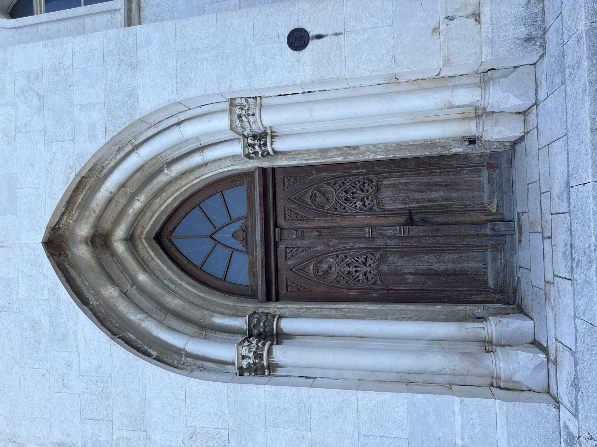 The image shows the entrance to the Cathedral of St. James in Šibenik, Croatia. The cathedral is a UNESCO World Heritage site and is known for its unique architecture, which combines elements of Gothic and Renaissance styles. The entrance features a large wooden door, arched stone doorway with intricate carvings, and a stained-glass window above the door. The cathedral is made of stone and is located in the city of Šibenik, Croatia. The image is taken from a low angle, looking up at the entrance.