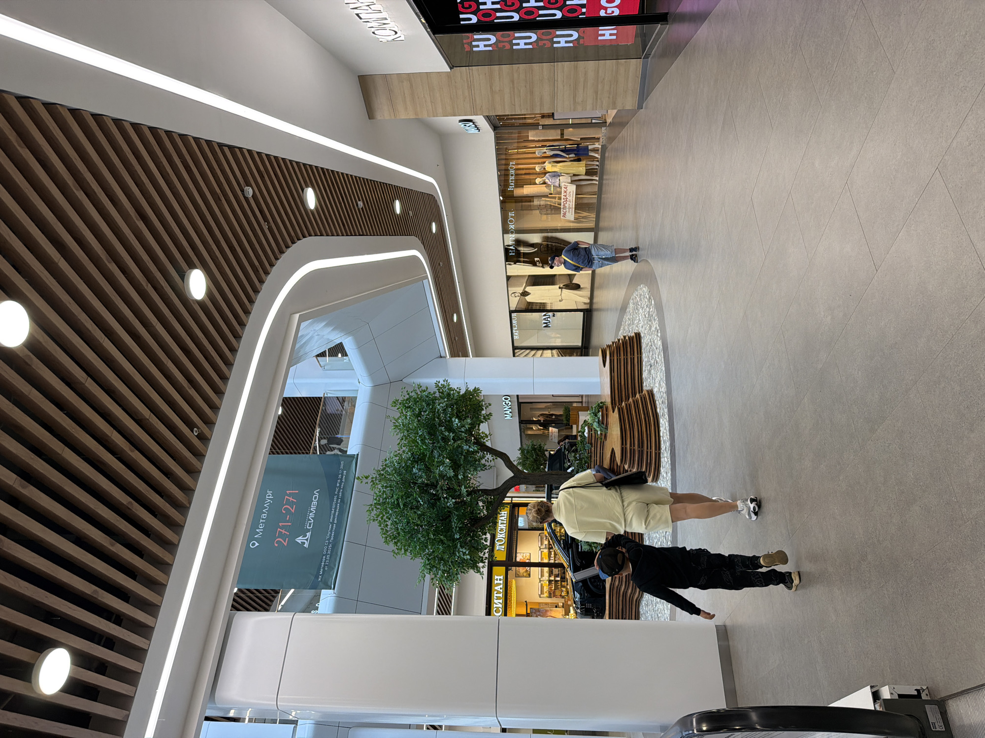 The image shows the interior of a modern shopping mall, likely in Izhevsk, Russia, as indicated by a banner that reads "Металлург 271-271" and a company name that appears to be "СИМВОЛ". In the foreground, a woman in a light-colored matching set is walking away from the camera, holding a dark bag over her shoulder. A young boy in a black hoodie and patterned pants walks ahead of her. They are on a wide, light-gray tiled floor. Behind them, a large, artificial tree with lush green foliage is situated in a circular raised bed made of stacked, curved wooden elements. A black car is parked behind the tree.
Further into the mall, several storefronts are visible. One store has a prominent yellow sign with Russian text, possibly reading "СИТАН" and "ТОКСИТАН". Another store has a "MANGO" sign. Beyond that, mannequins display clothing in store windows. A man in a blue t-shirt and shorts is standing near these windows. To the right, a store with a prominent red and white "HUGO" logo is visible. The ceiling is a distinctive feature, composed of parallel wooden slats with integrated lighting, and curved white architectural elements. The overall atmosphere is bright and contemporary.