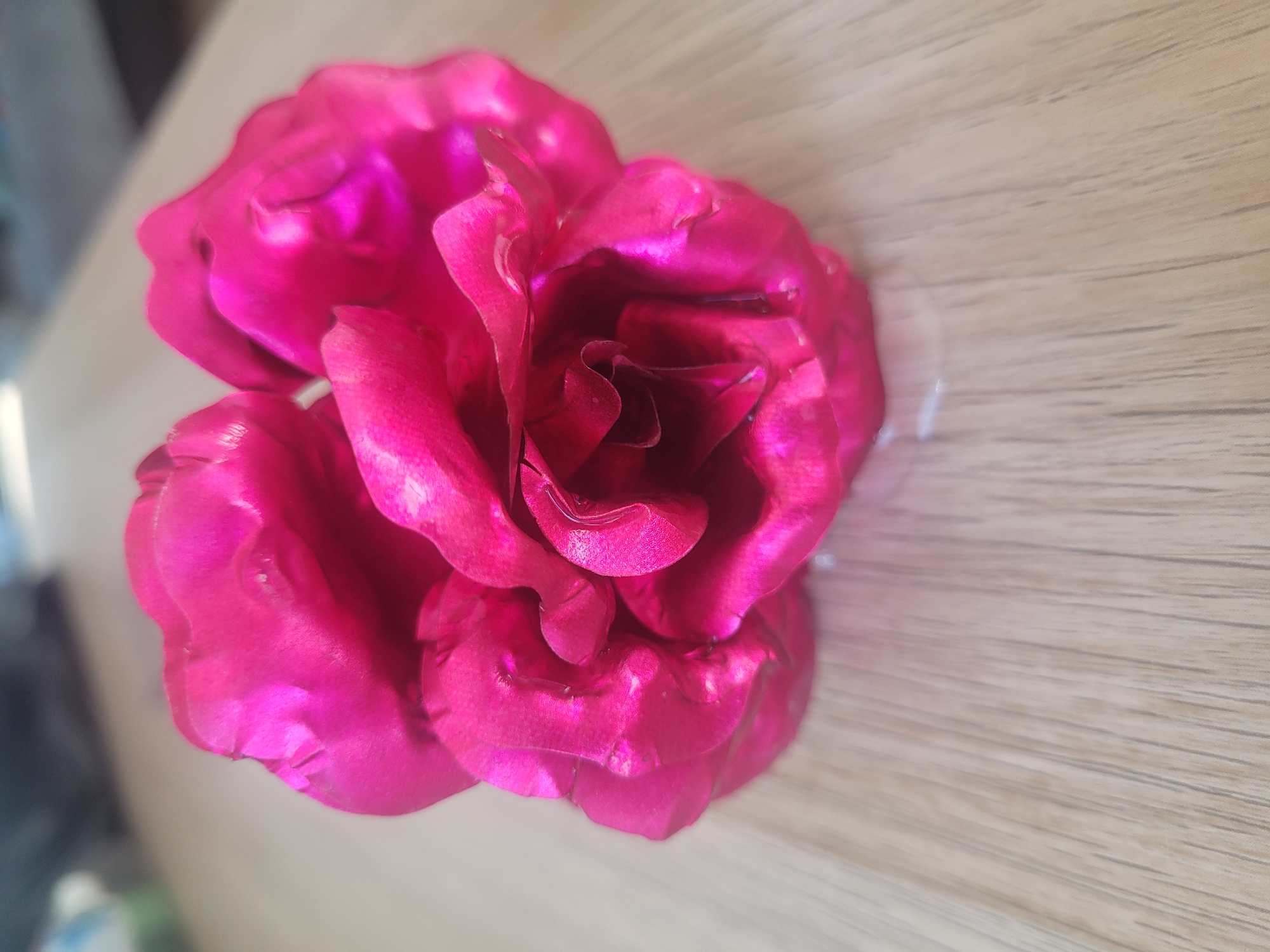 A close-up, top-down, slightly angled shot shows a bright pink, metallic-looking artificial rosebud resting on a light-colored, wood-grain textured surface. The rosebud is composed of several crinkled, overlapping petals that are highly reflective, capturing light and creating a glossy appearance. The surface beneath is out of focus, but appears to be a table or countertop with distinct wood grain lines running diagonally. The background is blurred and out of focus, with hints of a light-colored wall or curtain and a darker, possibly shadowed area. There are no people or animals visible in the image, and the setting appears to be indoors. The image is well-lit, with the primary light source appearing to be from the upper right, casting highlights and reflections on the rose. The overall impression is of a decorative object placed on a neutral surface.