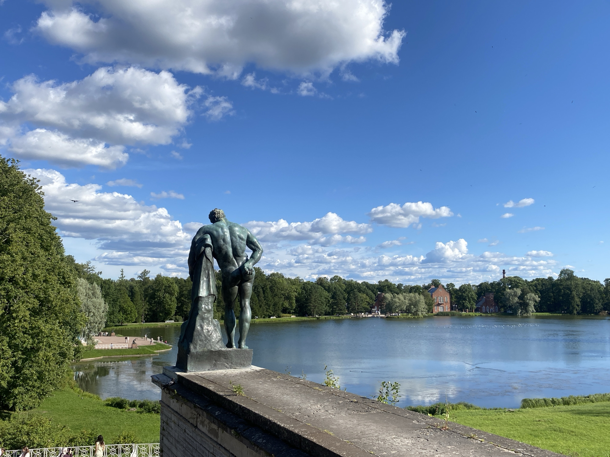 The image depicts a picturesque scene in Catherine Park, located in Pushkin, Russia, a suburban town south of St. Petersburg. In the foreground, a weathered stone balustrade runs along the edge of a raised platform. On this platform stands a bronze statue of a muscular, nude male figure, viewed from behind. He is draped with a cloth over his left shoulder and holds it with his left hand, while his right hand is placed on his hip. The statue is set against a backdrop of a tranquil lake.
The lake, reflecting the bright blue sky and scattered white clouds, stretches across the middle ground. On the far side of the lake, a lush green expanse of trees forms a dense backdrop, interspersed with several red-brick buildings, one of which has a prominent chimney. The buildings appear to be architectural elements of the park, possibly pavilions or other structures. Several small white birds are visible on the surface of the lake, and a few are flying in the distance.
To the left of the lake, a paved area with benches and people milling about is visible, bordered by a white decorative fence. The immediate surroundings of the lake are characterized by vibrant green grass. The sky above is a deep blue, adorned with fluffy white cumulus clouds. The overall atmosphere is serene and scenic, showcasing the natural beauty and architectural elegance of Catherine Park.