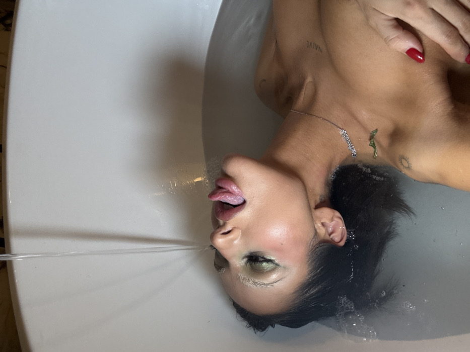 A woman reclines in a white bathtub filled with water. She tilts her head back, sticking her tongue out to catch a stream of water. She wears light-colored makeup and dark hair. A necklace and tattoos adorn her chest. The water stream is the main focal point, arcing towards her mouth. The image is taken from an overhead angle.