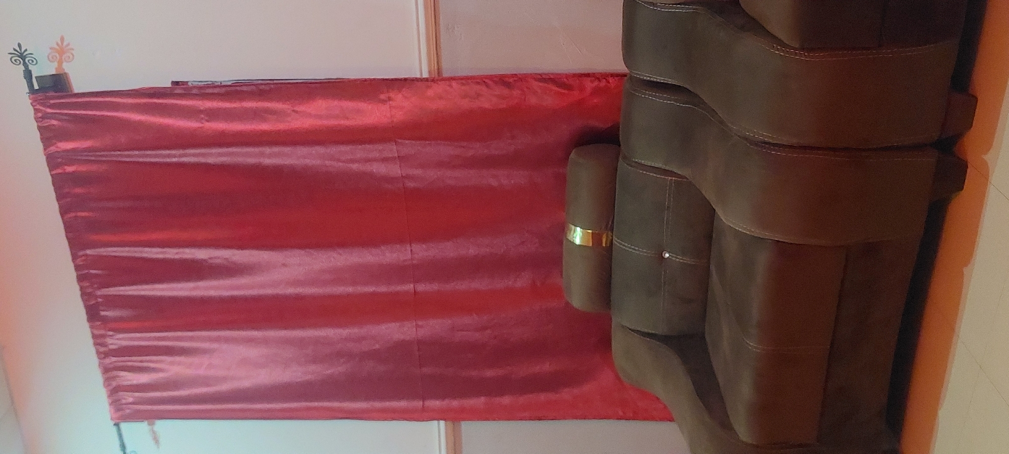 In an indoor setting, a plush, dark brown sectional sofa with white stitching is positioned in front of a vibrant red curtain. The sofa has multiple cushions, and a cylindrical bolster pillow with a gold band is placed on top of the back cushions. The curtain hangs from a decorative metal rod with ornate finials. The floor appears to be tiled with a light color, and a warm orange glow is reflected from the floor onto the bottom of the sofa. The room is dimly lit, suggesting it may be evening or the lights are off.