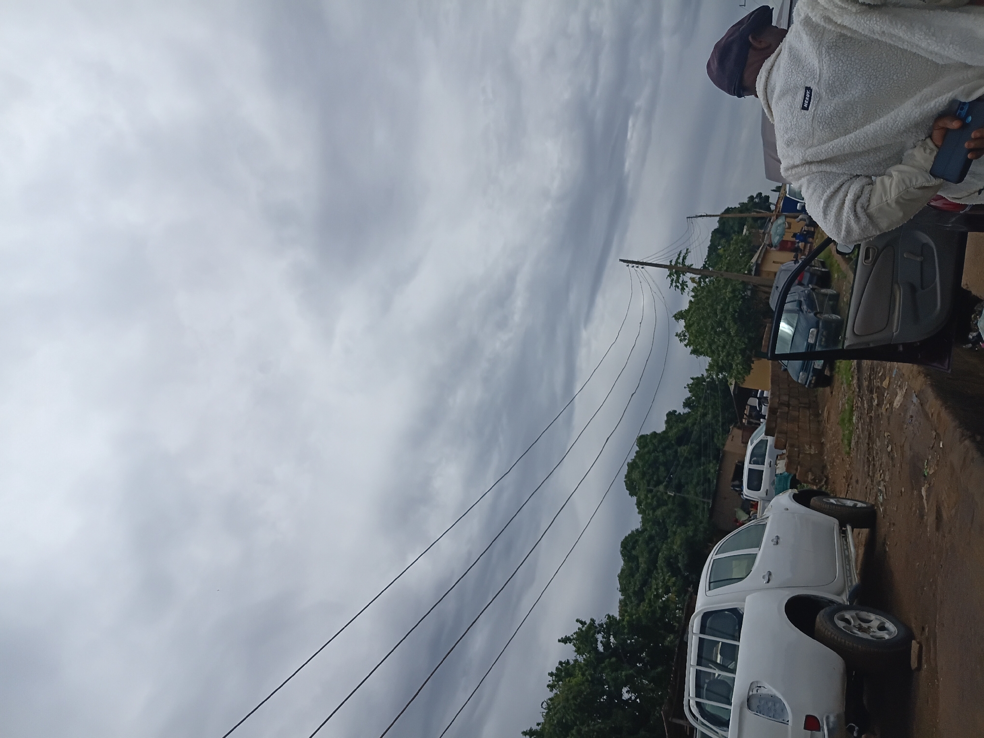 The image shows a cloudy sky with power lines stretching across the frame. In the foreground, a person wearing a beige sherpa jacket and a dark cap is visible from the back, holding a dark smartphone. To the left, a white pickup truck is parked on a dirt road. Several other vehicles, including a white van and a dark sedan, are also present. In the background, trees and buildings can be seen, suggesting an outdoor scene in a town. The overall setting appears to be a street or an informal parking area in Rafin Par, Nigeria, characterized by its unpaved roads and modest structures.