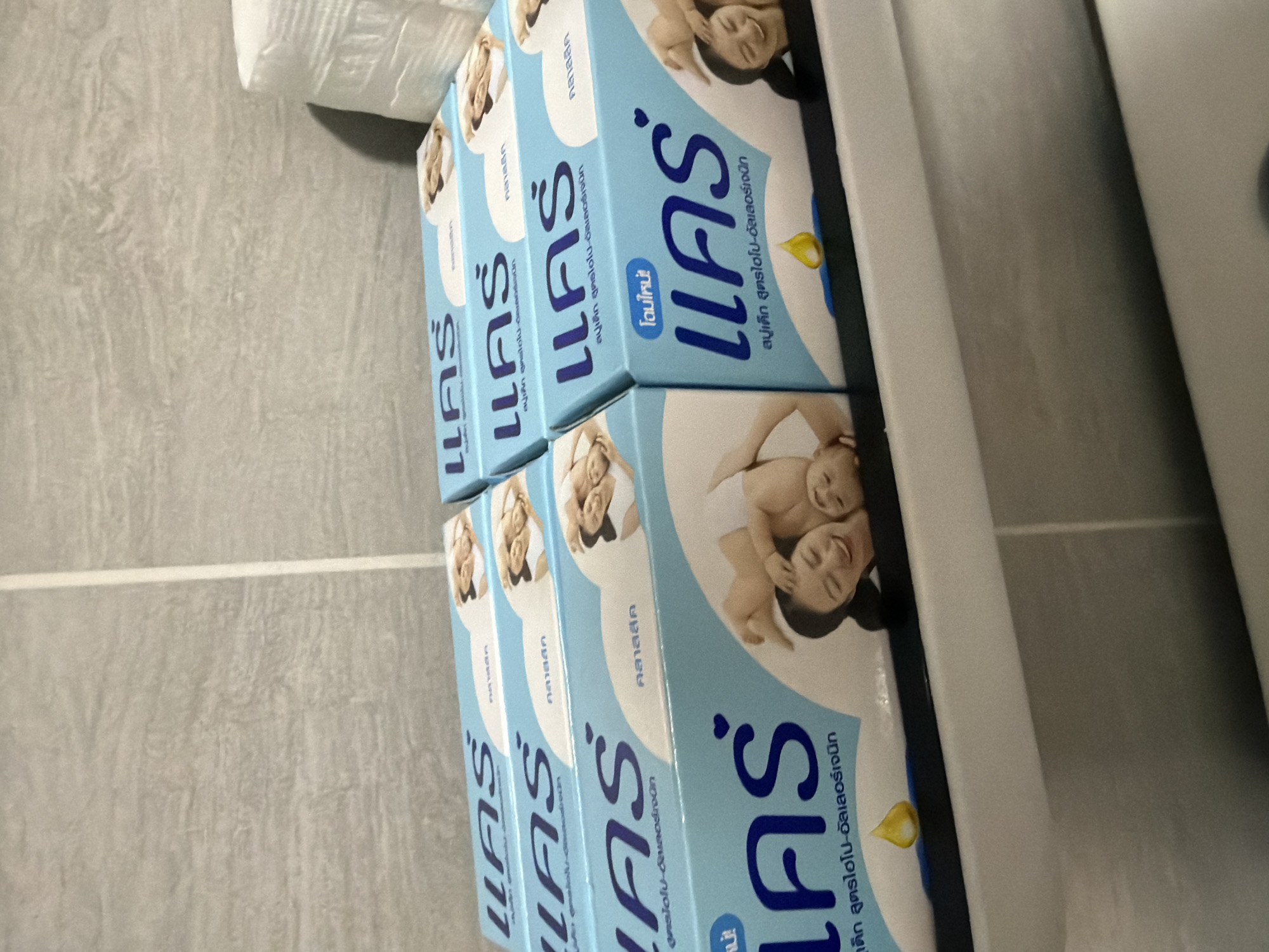 The image depicts six light blue cardboard boxes of "CARE" brand baby soap, stacked in two rows on a white shelf. Four boxes are in the front row, with two more partially visible behind them. Each box prominently features the brand name "แคร์" (CARE) in dark blue Thai script, often accompanied by a red heart icon. Additional Thai text on the packaging identifies the product as "สบู่เด็ก สูตรไฮโป-อัลเลอร์เจนิก" (Baby Soap, Hypo-allergenic formula), with some boxes indicating "คลาสสิค" (Classic) and one front-facing box stating "โฉมใหม่!" (New Look!). The graphic design on each box includes a smiling adult woman holding a smiling baby, positioned above a yellow oil droplet and a blue semicircular base. The background consists of a light grey tiled wall with a subtle, irregular pattern, and a distinct vertical white grout line on the right side. To the far right, a stack of white packaged paper products, resembling tissue or toilet paper, is partially visible. The scene is an indoor environment, likely a bathroom or storage area. The exclusive presence of Thai script on the product packaging situates the scene in Thailand. While specific Bangkok landmarks are not visible, this type of widely distributed consumer product is commonplace in urban centers throughout the country, including Bangkok.