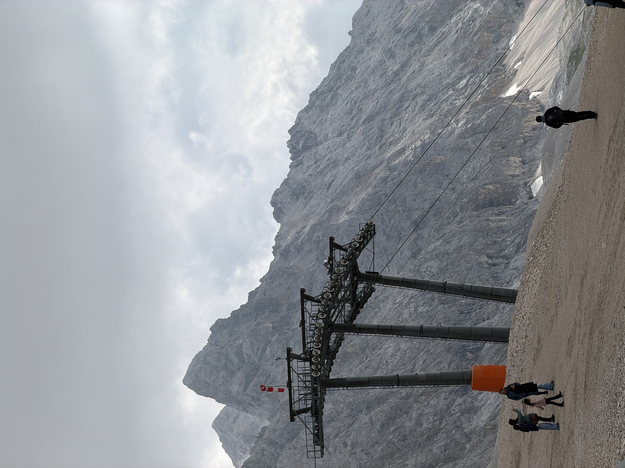 The image depicts the Zugspitze cable car station on a cloudy day, with the imposing Zugspitze mountain range serving as a dramatic backdrop. In the foreground, a group of tourists are visible on a gravel path, some taking pictures and others walking. A large orange cylindrical object stands near the cable car support structure. The cable car structure itself is a prominent feature, with multiple support pillars and a complex system of pulleys and cables. The mountain slopes are rugged and rocky, with patches of snow visible in some areas. The overall atmosphere is one of natural grandeur and human endeavor in a challenging mountain environment.