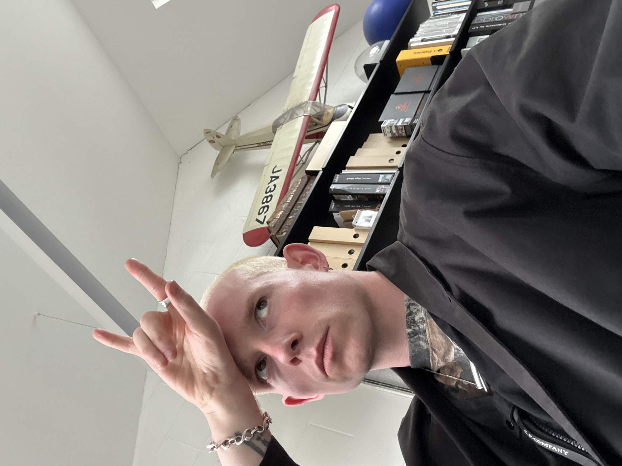 A low-angle selfie of a fair-skinned person with short, light-colored hair, wearing a dark jacket and a bracelet on their left wrist, is visible. They are making a hand gesture resembling "rock on" with their left hand. Behind them, a model airplane is mounted on the wall next to a bookshelf filled with books and objects. The setting appears to be indoors, with white walls and what appears to be overhead lighting. The name “C.P. Company” is visible on a portion of the jacket. There is no information present to indicate the location is the city of London, United Kingdom.