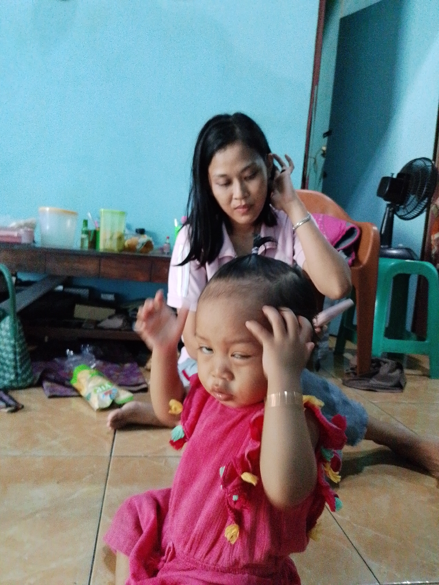 The scene is set indoors in Kecamatan Pati, Indonesia, evidenced by local packaging on the floor. A baby wearing a bright pink dress sits on a brown tiled floor, with her hands near her head. An adult woman with black hair is seated behind the baby. She is wearing a light pink shirt and appears to be touching her hair with her left hand, and holding a silver object in her right hand. In the background, there's a dark brown wooden table, a green plastic stool, and a black electric fan. A partially open door is visible in the background as well.