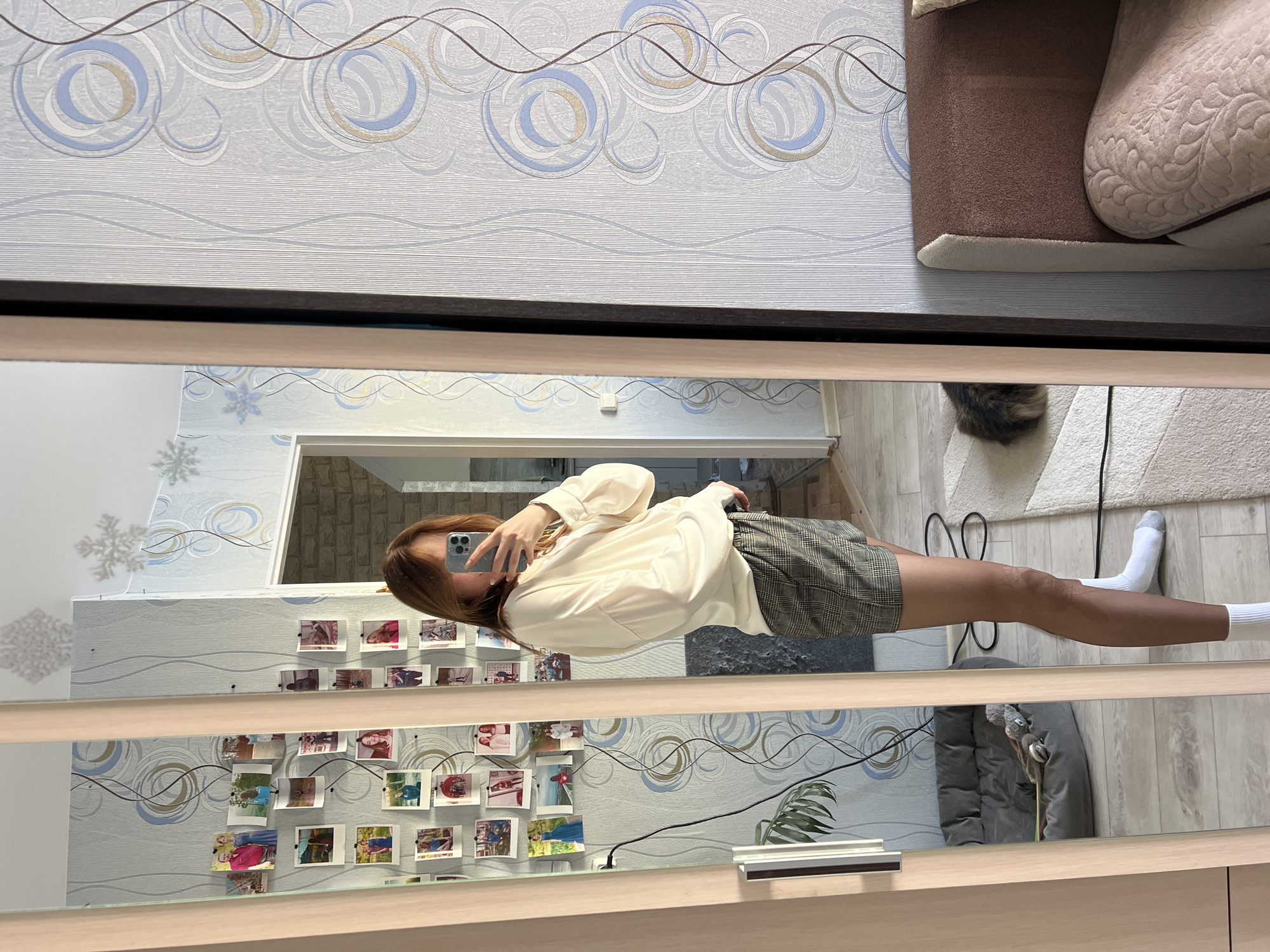 In an indoor domestic setting, identified as a location within the city of Chaikovsky, Russia, a young adult female with light brown hair stands sideways to a full-length mirror with a light wood frame, taking a selfie with a light-colored smartphone. She wears a loose-fitting cream long-sleeved shirt, grey and brown plaid shorts, and white crew socks. The mirror reflects a wall adorned with multiple small, unframed instant photographs clipped vertically, interspersed with white decorative snowflake decals on the upper portion. The wallpaper behind these decorations features a pattern of swirling blue, brown, and light-colored circles and wavy lines on a light grey background. In the lower left of the reflection, a grey pet bed containing a small toy is visible next to a green houseplant. A dark electrical cord runs across the light-colored laminate and cream shag rug flooring. A domestic cat with dark, fluffy fur is partially visible on the rug in the lower right portion of the reflection. To the right, outside the mirror's frame, a brown fabric sofa with a quilted cream armrest is partially visible. An open doorway reveals a room with light-colored brick-patterned walls in the background reflection.