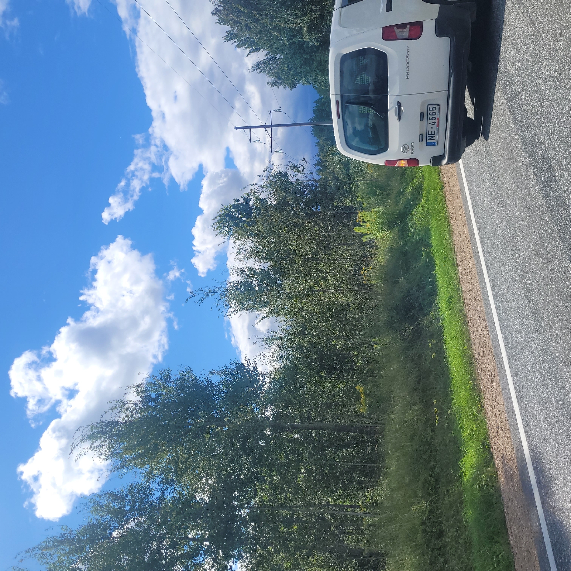 The image depicts a white Toyota Proace City van with the license plate NE-4665, traveling on an asphalt road. The van is seen from the rear, with the right side of the vehicle visible. The road has a white line marking on the right edge. To the left of the road, there is a grassy embankment with some vegetation and tall trees forming a dense line in the background. The sky above is blue with scattered white clouds, and a utility pole with power lines is visible in the distance, to the right of the trees. The scene appears to be a rural or semi-rural setting, possibly on the outskirts of the city of Ķekava, Latvia, given the license plate format. The lighting suggests it is daytime, likely during fair weather. There are no people visible in the image.