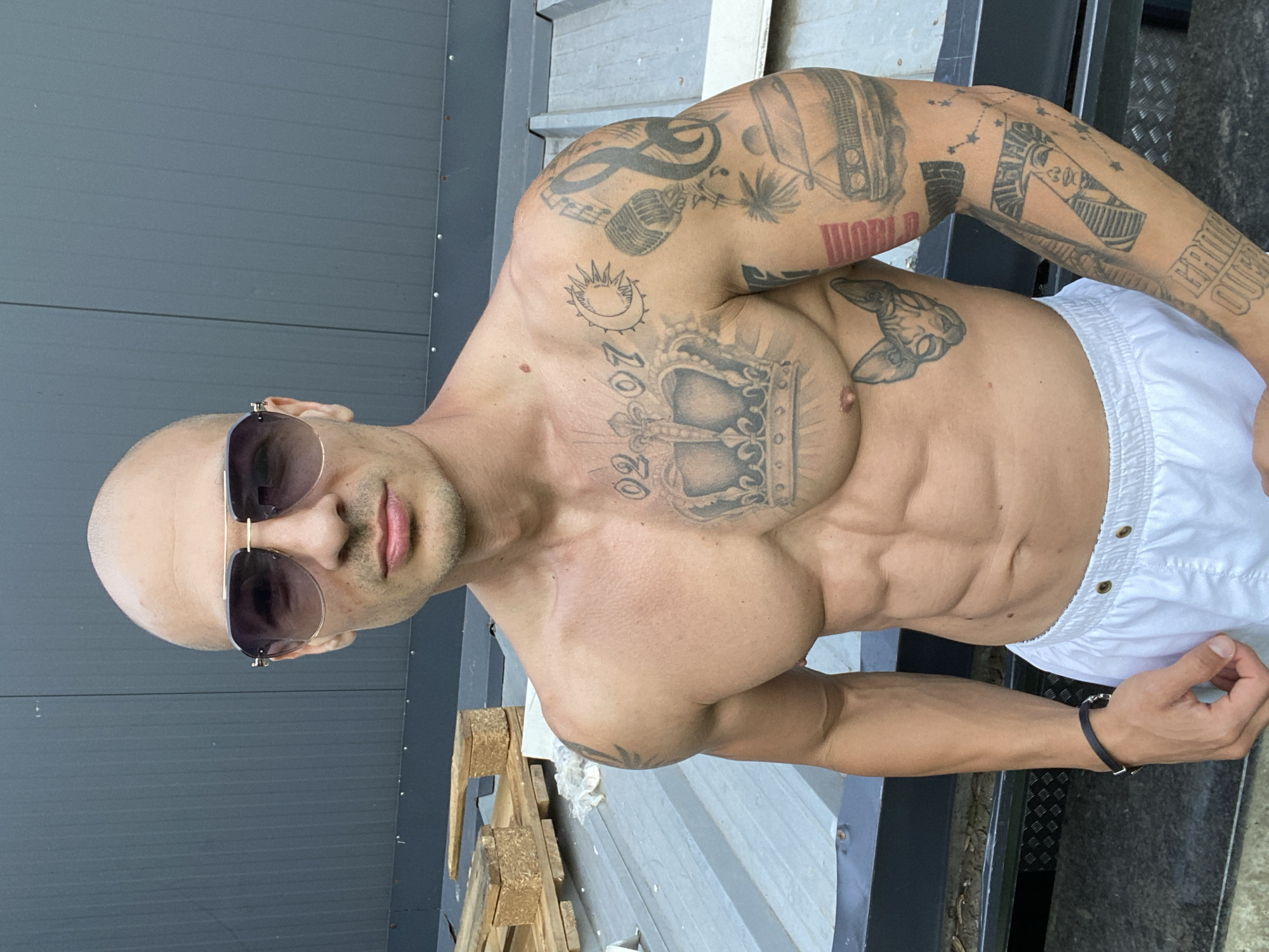 A bald, muscular man wearing aviator sunglasses and white shorts stands outdoors, his torso bare and covered in tattoos. He is positioned in front of a dark grey corrugated metal wall. To his left, several wooden pallets are stacked on a grey, metallic surface. His tattoos include a crown on his chest, a crescent moon and sun, a microphone, musical notes, a palm tree, a car, a cat's face, an Egyptian pharaoh's profile, and text. He is also wearing a black bracelet on his left wrist. The scene appears to be a rooftop or industrial area. There is no visual information to determine the specific location in Pleven, Bulgaria.