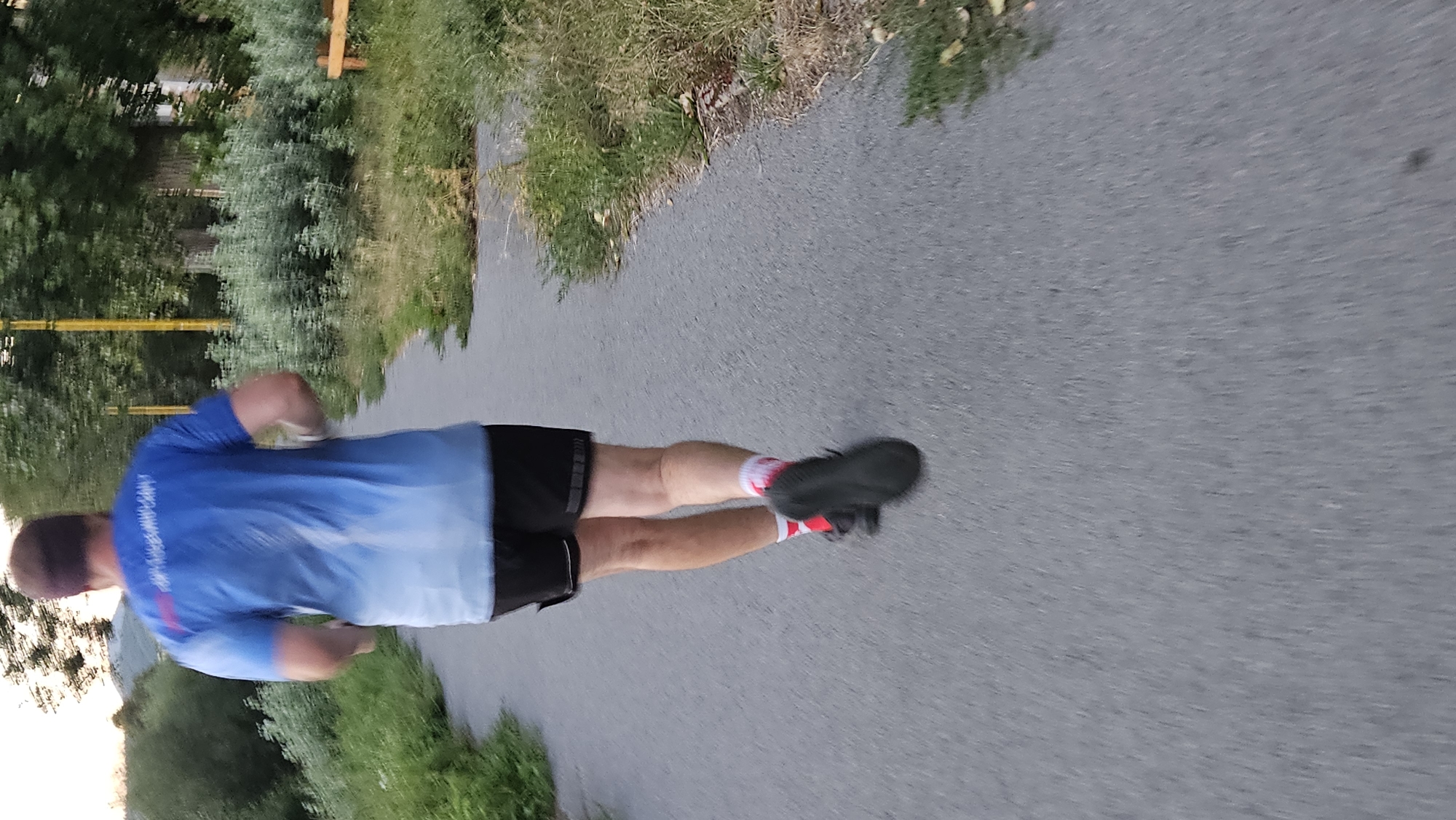 A person is captured from behind, running on a paved path. The runner is wearing a blue t-shirt with a gradient from dark blue at the top to white at the bottom, black shorts, red and white socks, and dark athletic shoes. The path appears to be in an outdoor, possibly park-like setting, with grass and trees visible on either side. In the background, there are trees, a yellow pole, and a wooden bench. The lighting suggests it might be late afternoon or early evening. The image's perspective is slightly downward and tilted, indicating it was taken by someone walking or running behind the subject. This scene could be located along the Sekčovský potok riverbank in Prešov, Slovakia, a common area for recreational activities.