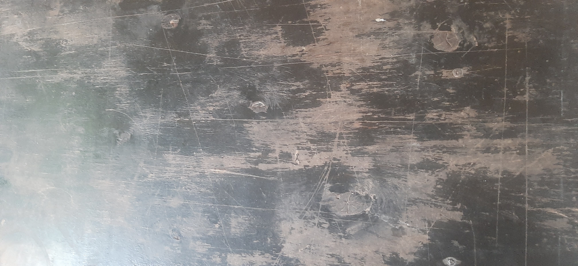 The image displays a close-up, overhead view of a dark, worn surface, likely a tabletop or floor. The surface is heavily scratched, with lighter colored abrasions and scuffs crisscrossing the dark background. Several circular indentations or bumps are visible, some appearing to be screw heads or remnants of something attached to the surface. Faint lines, possibly from the grain of the material or previous markings, are also present. The lighting creates a somewhat mottled appearance, with areas of lighter reflection. There are no visible people, animals, or distinct objects in the frame. The overall impression is one of an aged and heavily used surface. The location context provided is Yessentukskaya, Russia, but this information does not offer visual cues within the image itself. There is no discernible text.