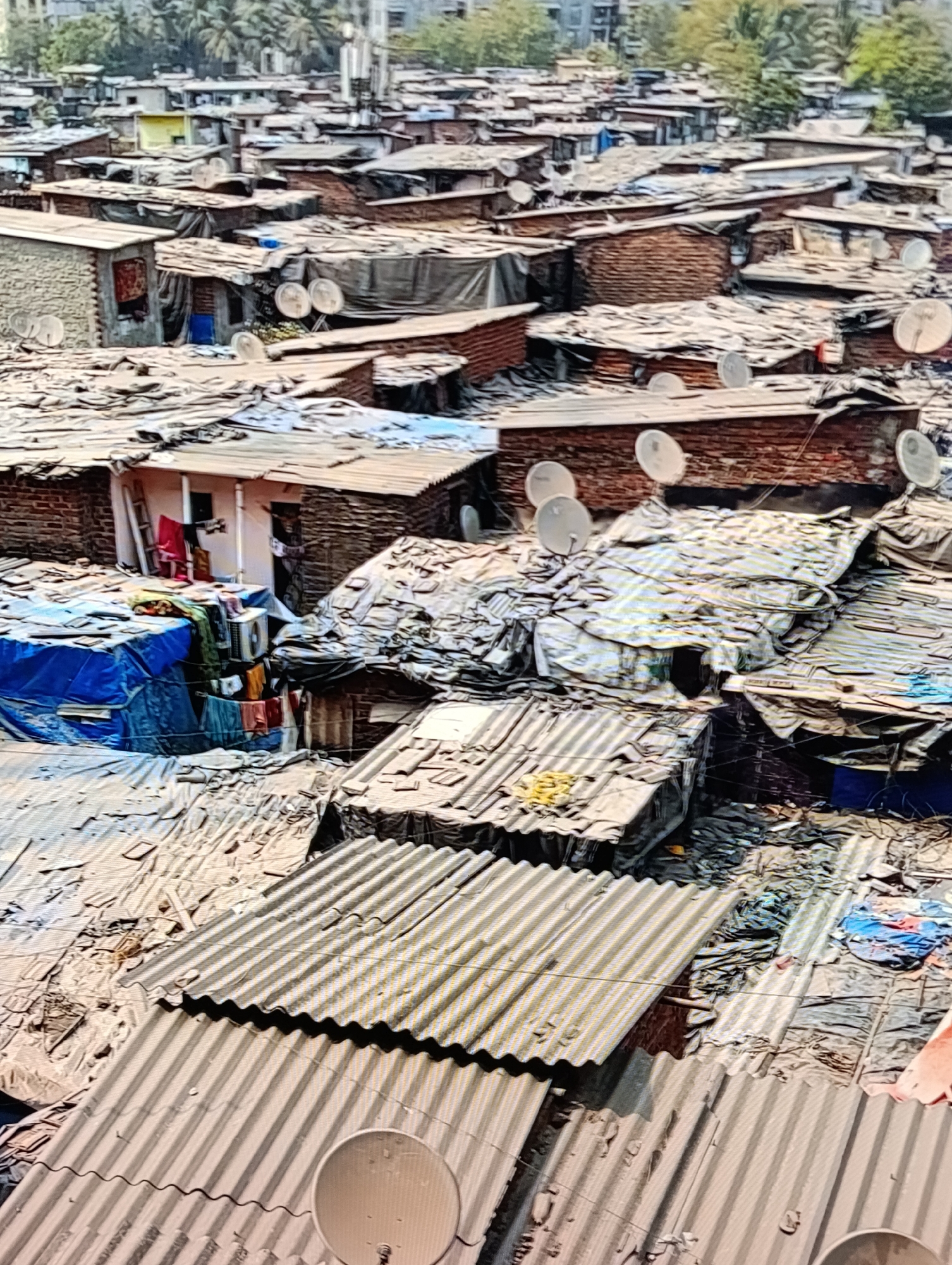 The image depicts a densely packed slum in an unknown location. Numerous small, makeshift dwellings are constructed from brick, corrugated metal, tarpaulins, and other found materials. Many roofs are covered with satellite dishes. Laundry is hanging outside some of the structures. The environment appears impoverished. There is no explicit interaction among people in the image. In the background, buildings and trees can be seen.