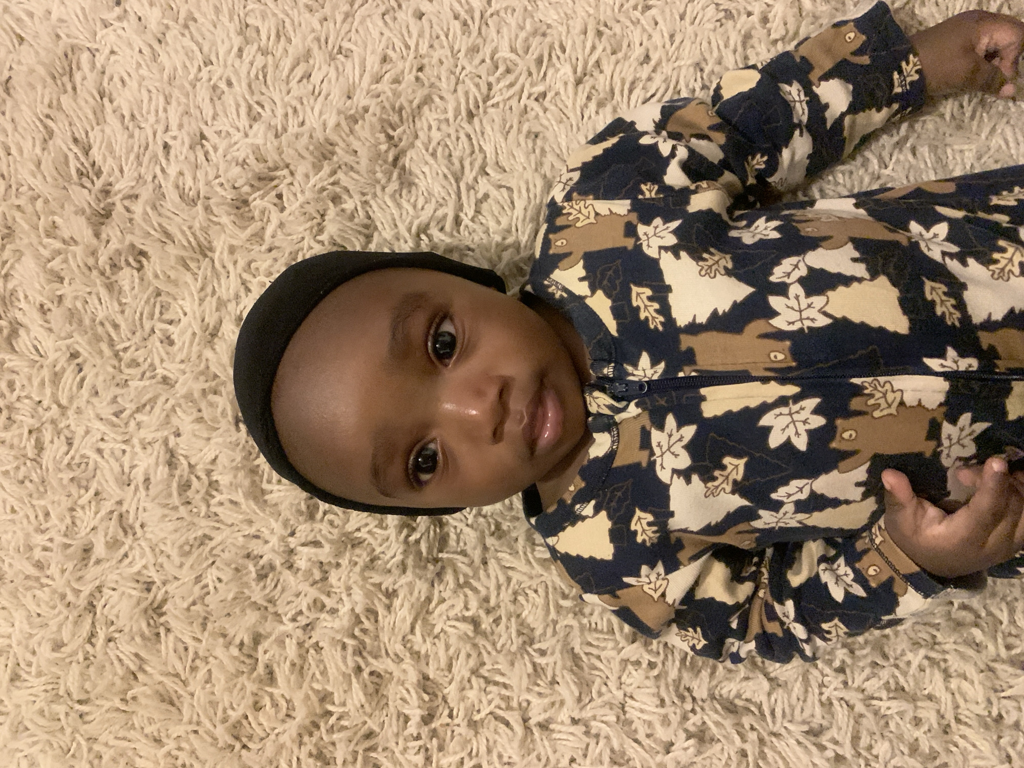 A baby wearing a black cap and a blue, brown, and white patterned jumpsuit is lying on a beige shag rug. No information is available about the location in Barakin Akawo, Nigeria.
