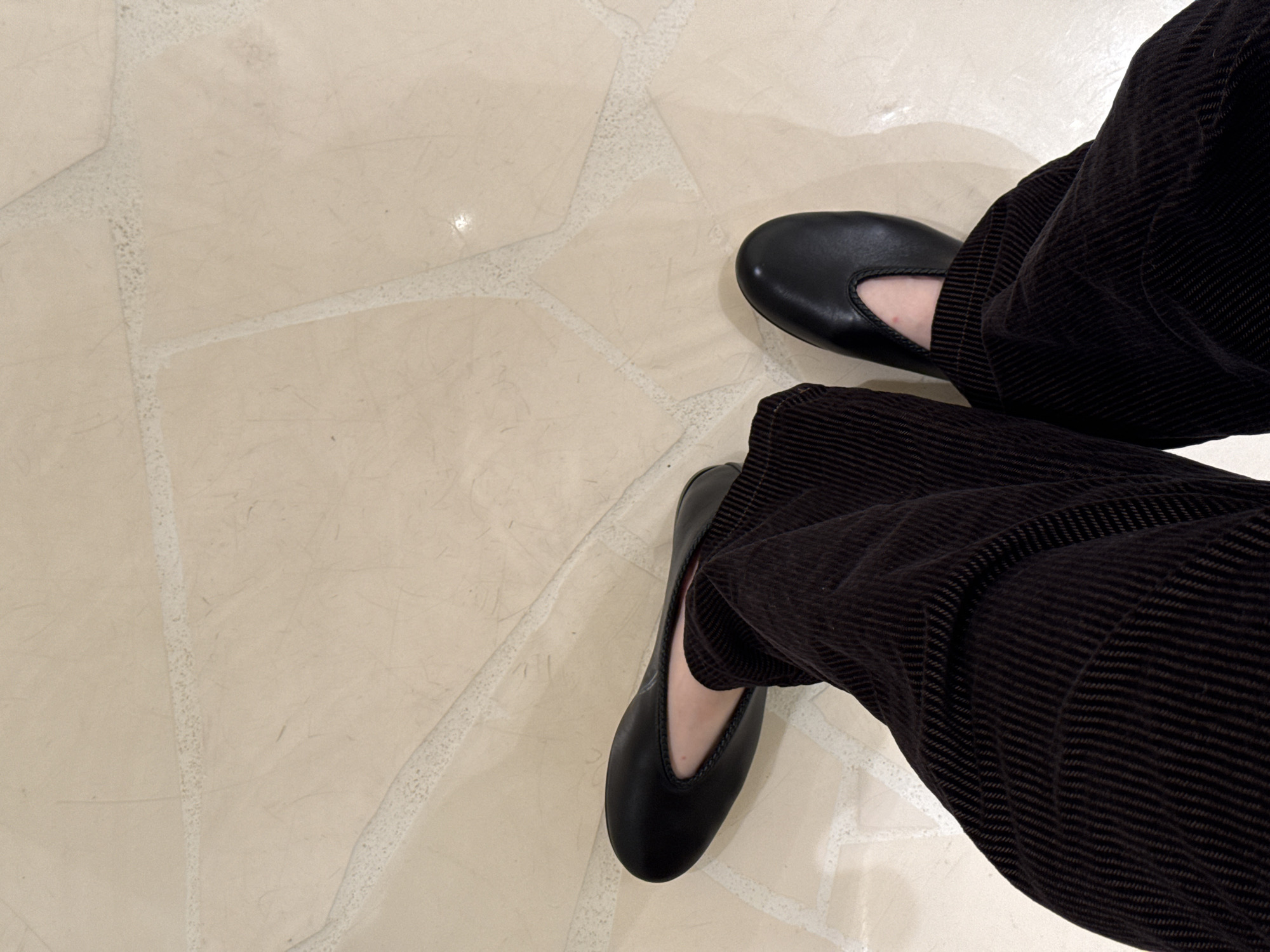 A person wearing black corduroy pants and black ballet flats stands on a light-colored tiled floor. The tiles are a cream color with irregular shapes and light grout lines. The person's feet are positioned near the right side of the image, with the left foot closer to the viewer. The pants are slightly bunched around the ankles. No other people or objects are visible. There is no information to indicate this image was taken in the city of San Diego.