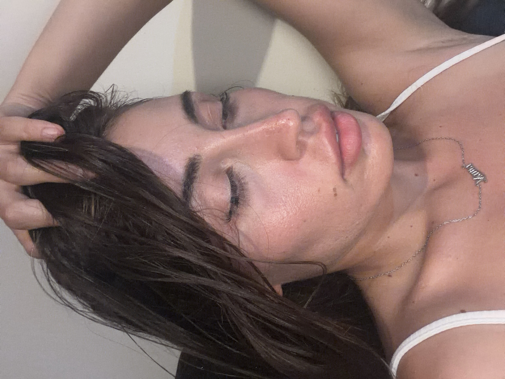 The image shows a close-up of a person's face, tilted downwards and to the left, with their hand pulling at their hair. The person has dark hair, is wearing a white top, and a silver necklace with a pendant that appears to spell "Mama". The lighting is soft and creates a glow on the skin. The background is a plain, light-colored wall. There are no discernible elements in the image that would allow for the identification of the location as Tersefanou, Cyprus.