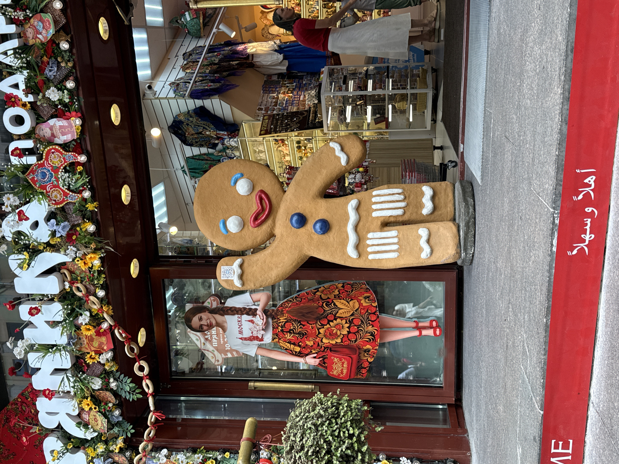 This image captures the exterior of a shop in Moscow, Russia, likely a souvenir or gift shop given the decorative elements. In the foreground, a large, stylized gingerbread man figure stands to the right, partially obscuring a life-sized cardboard cutout of a woman on the left. The woman is depicted wearing a white t-shirt with "МОСКВА" (Moscow) written on it and a traditional Russian folk-patterned skirt, holding a red handbag.
Above the shop entrance, large white lettering, possibly part of the shop's name, is decorated with garlands of artificial flowers, fruits, and small Russian nesting dolls (matryoshka dolls). A string of dried bread rings, a traditional Russian decoration, is also visible.
The shop's entrance is a glass door, revealing shelves stocked with various goods, including scarves and souvenirs, inside. A woman in a red top and a long white skirt is seen inside the shop, interacting with a child. The shop floor is covered with dark and light grey paving stones. A red band with white lettering, containing both English ("ME") and Arabic ("أهلا وسهلا" - Welcome), runs along the bottom of the image, suggesting a welcoming message to international visitors. The overall impression is a festive and culturally themed retail establishment in Moscow.