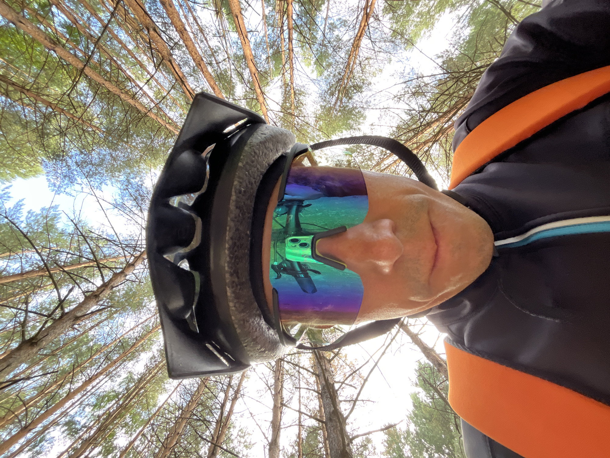 This is a first-person perspective, upside-down selfie taken by a person wearing a black bicycle helmet and multi-colored cycling glasses. The subject is a person with a visible portion of their face, including their nose and mouth, visible. They are wearing a dark jacket with bright orange and blue accents.
The background of the image is dominated by the canopy of a pine forest, with tall trees reaching upwards towards a bright, overcast sky. The perspective is looking directly up through the branches, creating a sense of being immersed in the forest. The lighting suggests it is daytime, likely mid-morning or afternoon, with diffused light filtering through the trees.
Reflected in the subject's cycling glasses, a smartphone can be seen, displaying a green interface with what appears to be camera controls or an app. The reflection also shows the surrounding environment and the person's own helmet and face from a different angle.
The overall impression is of someone enjoying an outdoor activity, possibly mountain biking, given the attire and helmet. The setting is a natural, wooded environment, and the mood seems to be one of active recreation. No visible text is present in the image. The location context provided is Chaikovsky, Russia.