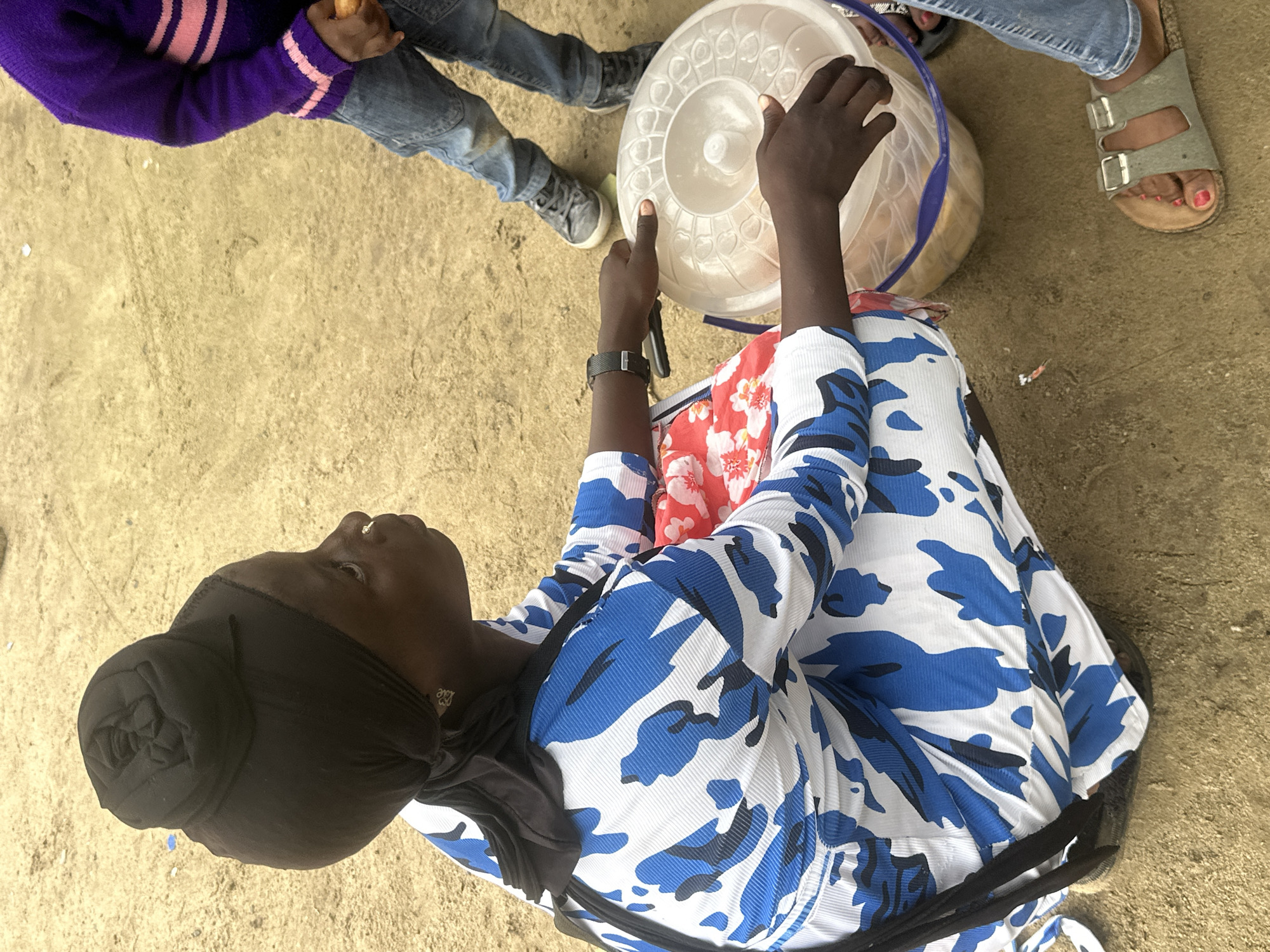 In Ganyingbo, Nigeria, a woman is kneeling on sandy ground, facing right in profile. She wears a black hijab styled into a bun and a white and blue camouflage-patterned long-sleeved dress. Her left hand rests on a clear plastic container with a blue rim, filled with food items. A child in a purple sweater with pink stripes and blue jeans stands to her right, holding an orange food item. Another person's legs and feet, wearing blue jeans and sandals, are visible in the foreground and to the right. The environment is dusty and appears to be an outdoor market or communal gathering space.