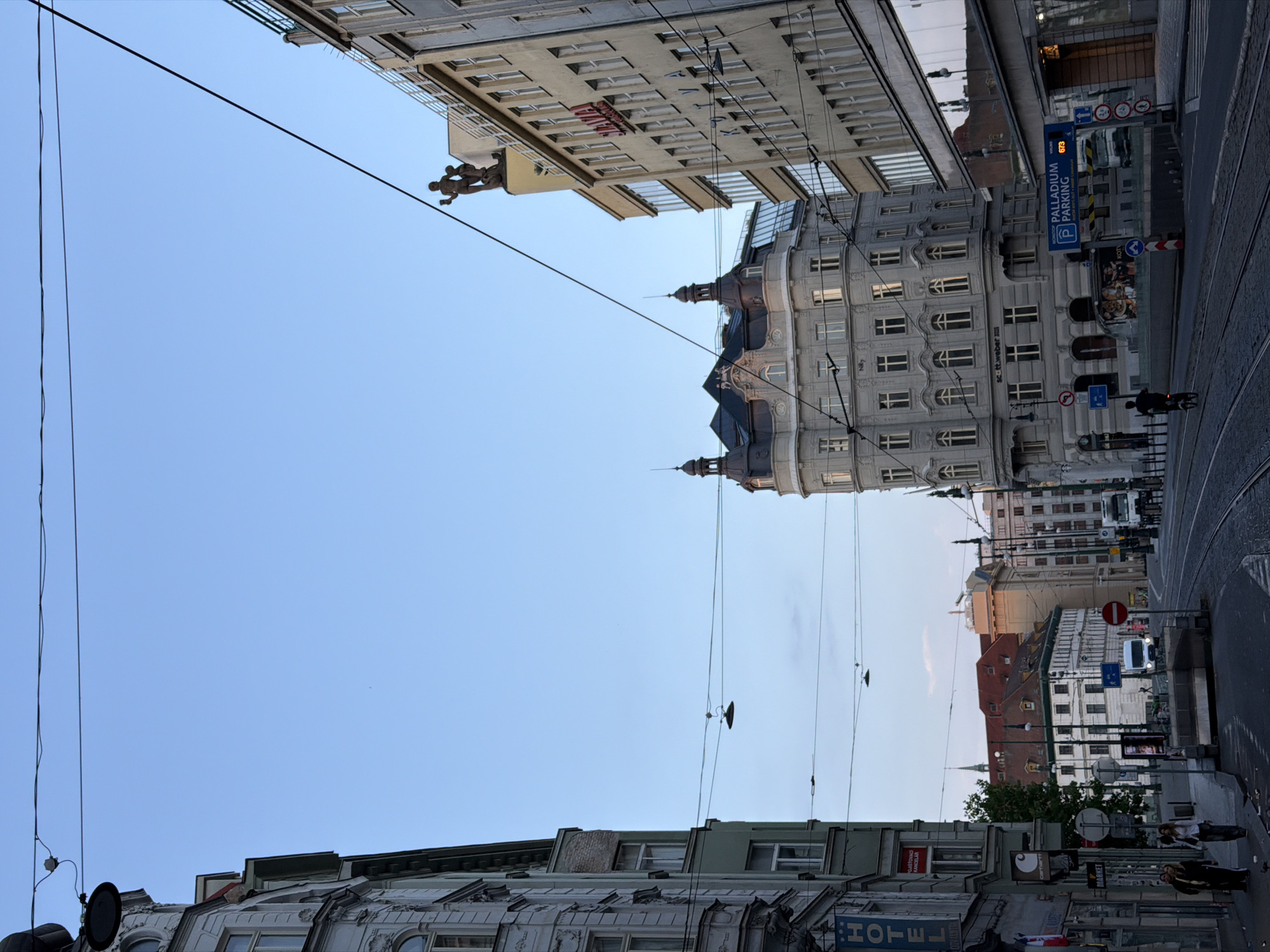The image captures a street scene in Prague, Czechia, likely near Palladium shopping center, given the prominent "PALLADIUM PARKING" sign. The photograph is taken from a street level perspective, looking up a wide street with tram tracks. The sky is a clear, pale blue, indicating it is either early morning or late afternoon.
On the left side of the street, a building with a prominent "HOTEL" sign is visible, featuring ornate architectural details. Further down the street on the left, there are shops and people walking.
In the center of the image, a distinctive building with rounded corners and multiple floors, featuring numerous windows, stands out. The architecture suggests a historical European style.
To the right, a taller, modern-looking building with a red "SLAVIA" sign is depicted. Above this building, a statue of two figures, likely a mythological or allegorical representation, is positioned on a pedestal.
Further down the street, traffic can be seen, including a white van and a truck, and tram tracks run across the road. There are various traffic signs and street lights present. Overhead, power lines crisscross the sky. A person on a bicycle is visible riding down the street. The overall impression is of a busy urban environment with a mix of historical and modern architecture.