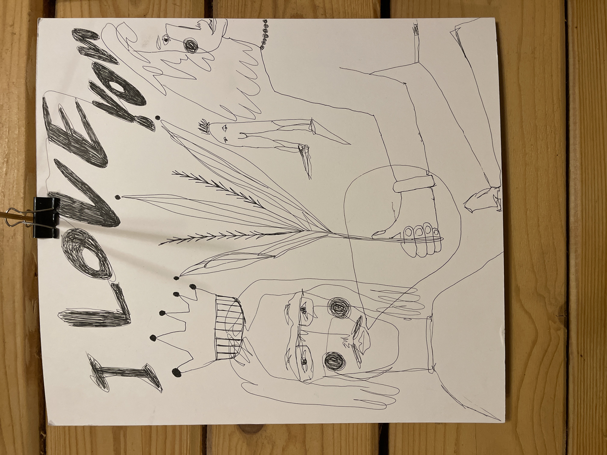 The image features a continuous-line drawing on a white sheet of paper, fastened by a black binder clip to a light brown wooden surface. The photograph shows the paper rotated 90 degrees clockwise, but the drawing's intended orientation is vertical.
The artwork depicts two prominent figures and a smaller one. On the lower portion of the drawing, a figure, likely a woman, is shown wearing a crown. Her face is detailed with large, circular eyes, a nose, and a mouth, and she has long hair. Her expression is somewhat pensive. Her left hand is visible, reaching towards the center, where it holds a cluster of thin plant stalks resembling wheat or palm fronds.
On the upper right side, a second, more abstract and elongated figure is drawn. This figure also appears to be a woman, characterized by long, wavy hair and a simple beaded necklace. Her face is minimally rendered with circular eyes and a small mouth. A small, stick-figure-like person stands between the two larger figures.
Along the left edge of the paper, the words "I LOVE YOU" are written vertically in bold, block letters, filled with scribbled lines. The drawing style is simple, abstract, and gives a personal, heartfelt impression.