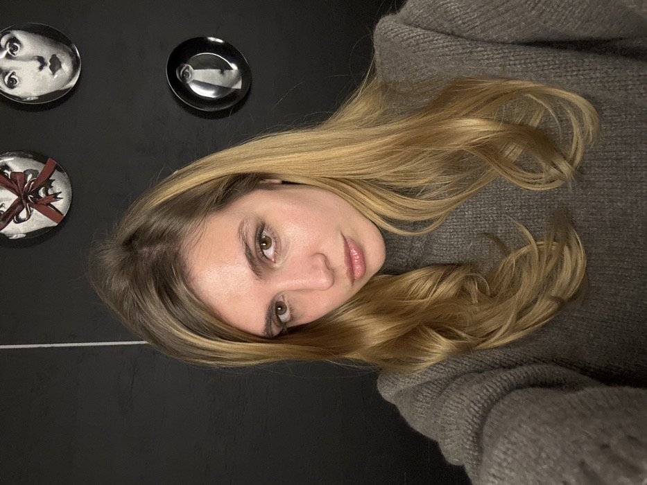 A woman with light brown eyes and blonde hair is wearing a gray sweater. She is looking at the camera with a slight tilt of her head. Behind her, there's a black surface with three decorative plates affixed to it. One plate features a face, another is plain, and the third is decorated with a red bow. A white line appears in the lower-left corner of the image.