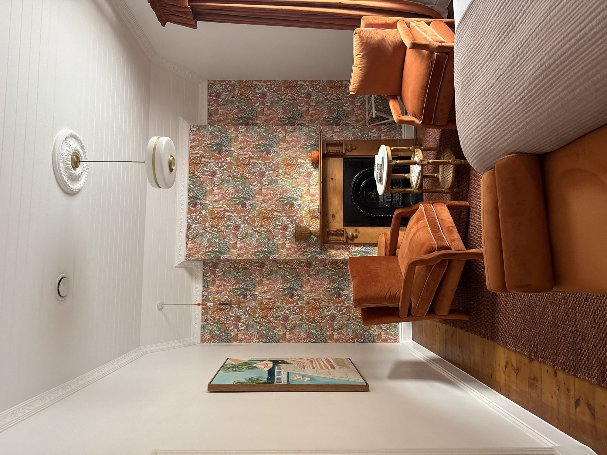 The image shows an interior space, with a focus on design elements. Two orange armchairs with white trim are placed near a wooden fireplace against a wall covered in a floral, vintage-style wallpaper. A small, ornate table with a white top sits between the chairs and the fireplace. Above the table and behind the right armchair is a large piece of furniture, possibly a sofa or bed. On the wall opposite the fireplace, a framed painting hangs. The wall is white with vertical panels and decorative molding. A modern wall sconce is mounted on the wall. The floor is a combination of hardwood and a brown area rug. The space appears to be in a residential setting. There is no visible evidence to suggest this location is in Cape Town, South Africa.