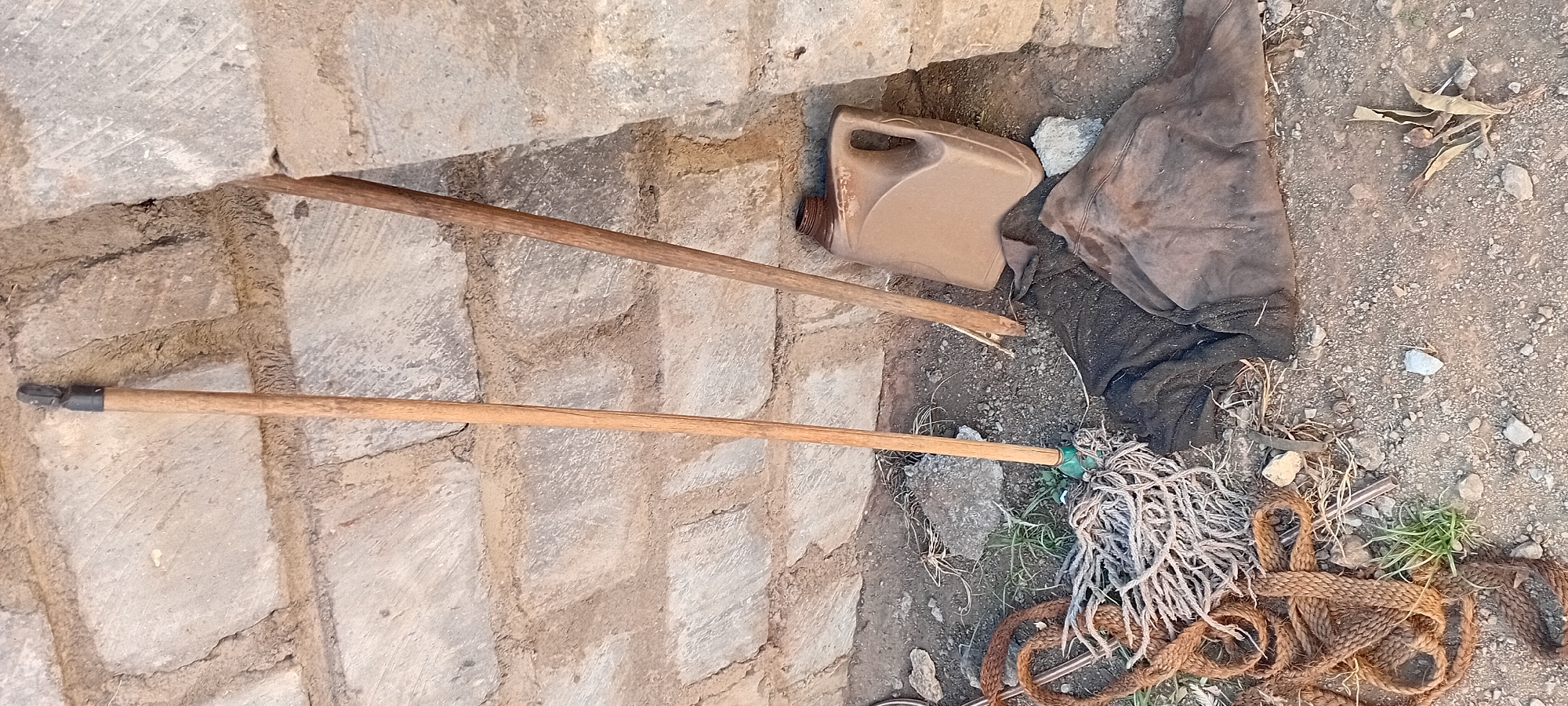 Two wooden poles, one with a mop head attached and another leaning against a brick wall, are positioned on the ground next to a brown plastic jug. Several pieces of rope and what appear to be rags or fabric are also present on the ground. The background consists of a rough brick wall. The scene is located in Kijabe ward, Kenya.