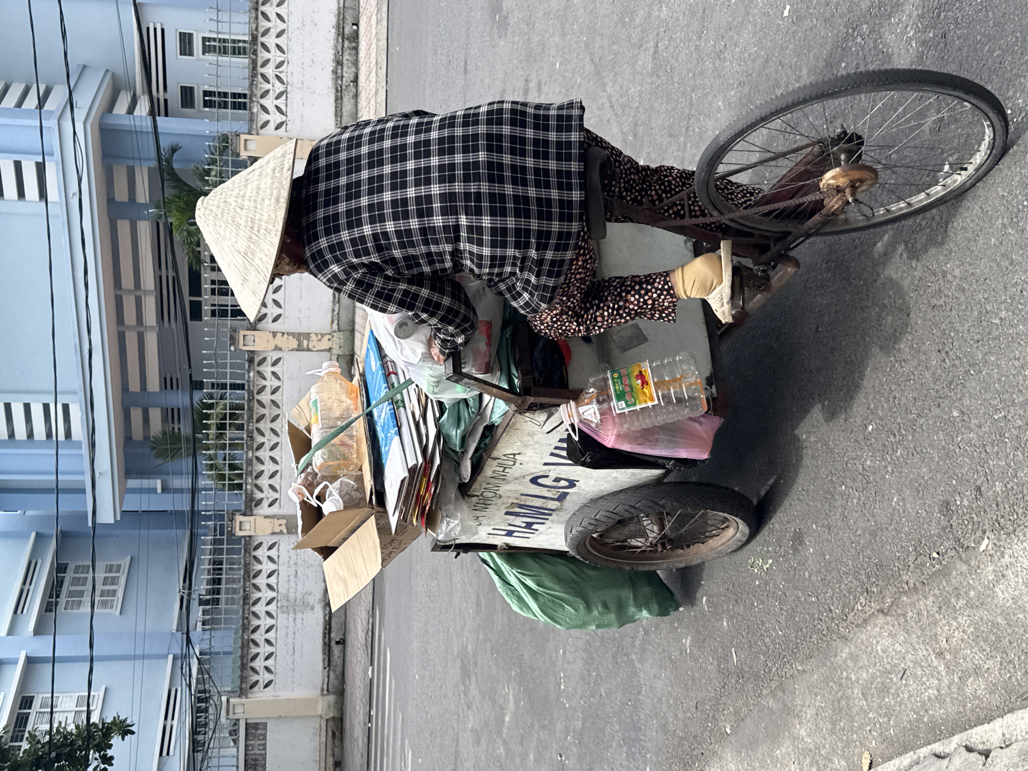 An individual wearing a conical hat and a black and white plaid shirt rides a tricycle laden with recyclables. The tricycle is a weathered brown metal frame with a single wheel in the front and two in the back. The individual is seated and appears to be pedaling. The cargo consists of cardboard boxes, plastic bottles, and green plastic bags. Text is visible on the side of the tricycle, appearing to be in Vietnamese. The background shows a light blue building with white shutters and a white fence with patterned cutouts. The scene is on an asphalt street, suggesting an urban environment. The image was taken in Nha Trang, Vietnam.