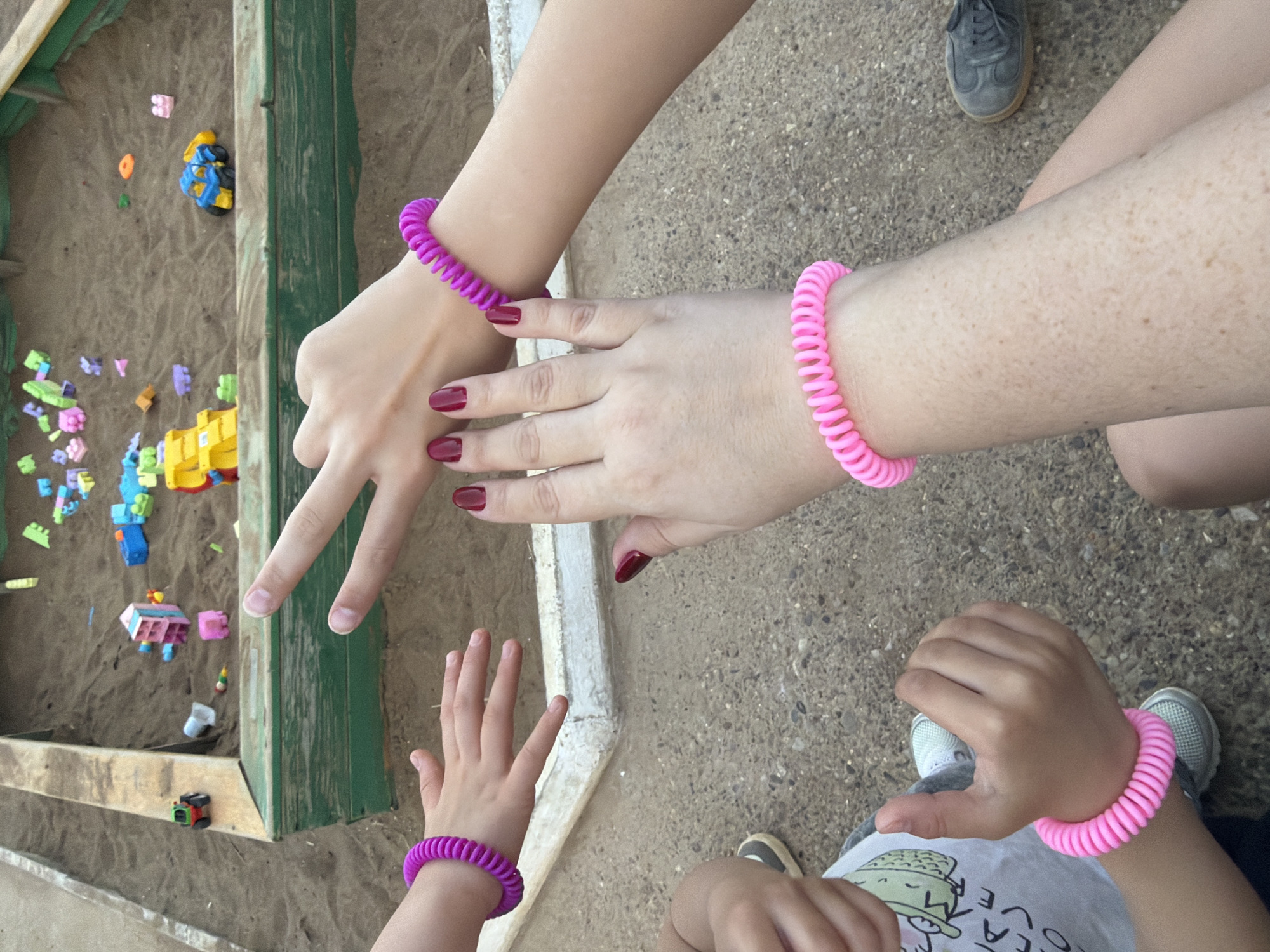 The image shows a sandbox with scattered colorful plastic toys. An adult's hand with red nail polish and a pink spiral bracelet points towards the sandbox. A child's hand with a matching purple bracelet reaches toward the adult's hand. The ground appears to be concrete or paved. Another adult leg and shoe are visible, also with a pink bracelet, as well as the torso and arm of a child. The location is unidentified.