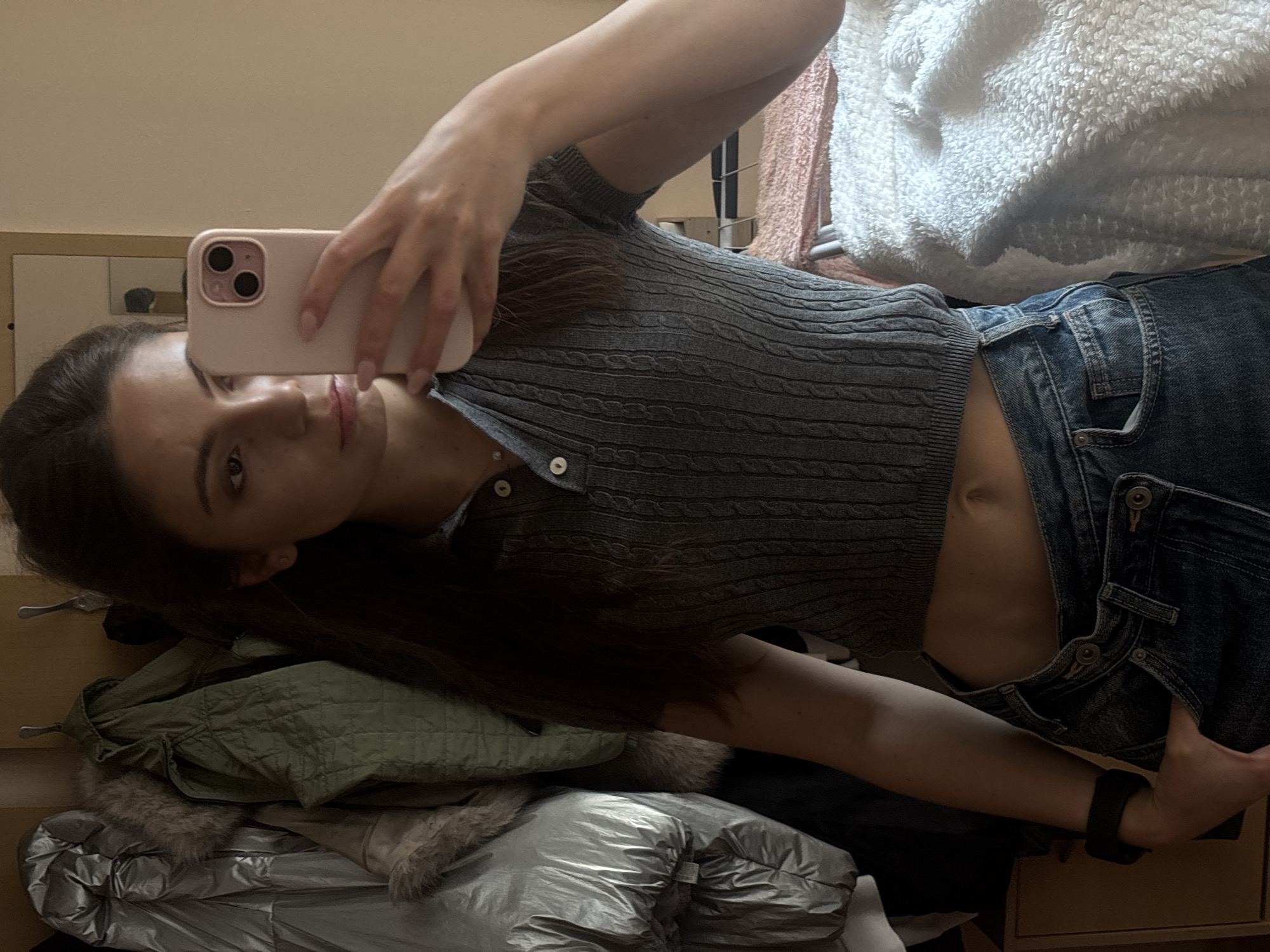 This is a self-portrait taken by a young woman in what appears to be a bedroom in Varna, Bulgaria. The woman is holding a pink smartphone in her right hand, angled to capture her reflection in a mirror, which is implied to be to her left and out of frame. She is wearing a grey, cable-knit, short-sleeved crop top with a buttoned collar and blue denim jeans, unbuttoned and partially unzipped. Her long, dark hair is visible, falling around her shoulders and face.
The background is cluttered with various items, suggesting a lived-in space. To the woman's left and below her, there are several pieces of clothing, including a green quilted jacket with fur trim and a shiny, silver puffer jacket. Behind her and to the right, a fluffy white blanket and a pink towel are visible, draped over what seems to be a rack or furniture. The overall lighting suggests it could be daytime, with the soft lighting possibly coming from a window or diffused artificial light. The woman's expression is neutral, and she appears to be casually posing for the photo. There is no visible text in the image.