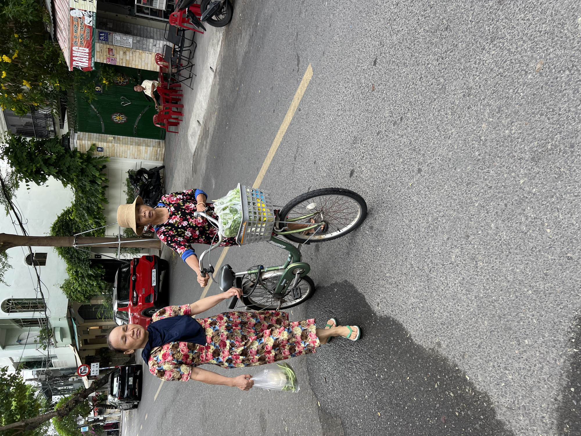 In Nha Trang, Vietnam, two women are walking on a paved street. The woman on the left, wearing a floral dress and a dark blue scarf, carries a plastic bag filled with produce. The woman on the right, wearing a floral blouse and a straw hat, pushes a green bicycle with a basket containing vegetables. The street is lined with buildings, trees, and parked vehicles. A red SUV is parked on the right side of the street, and a black car is visible on the left. The scene appears to be a typical street view in a Vietnamese city.