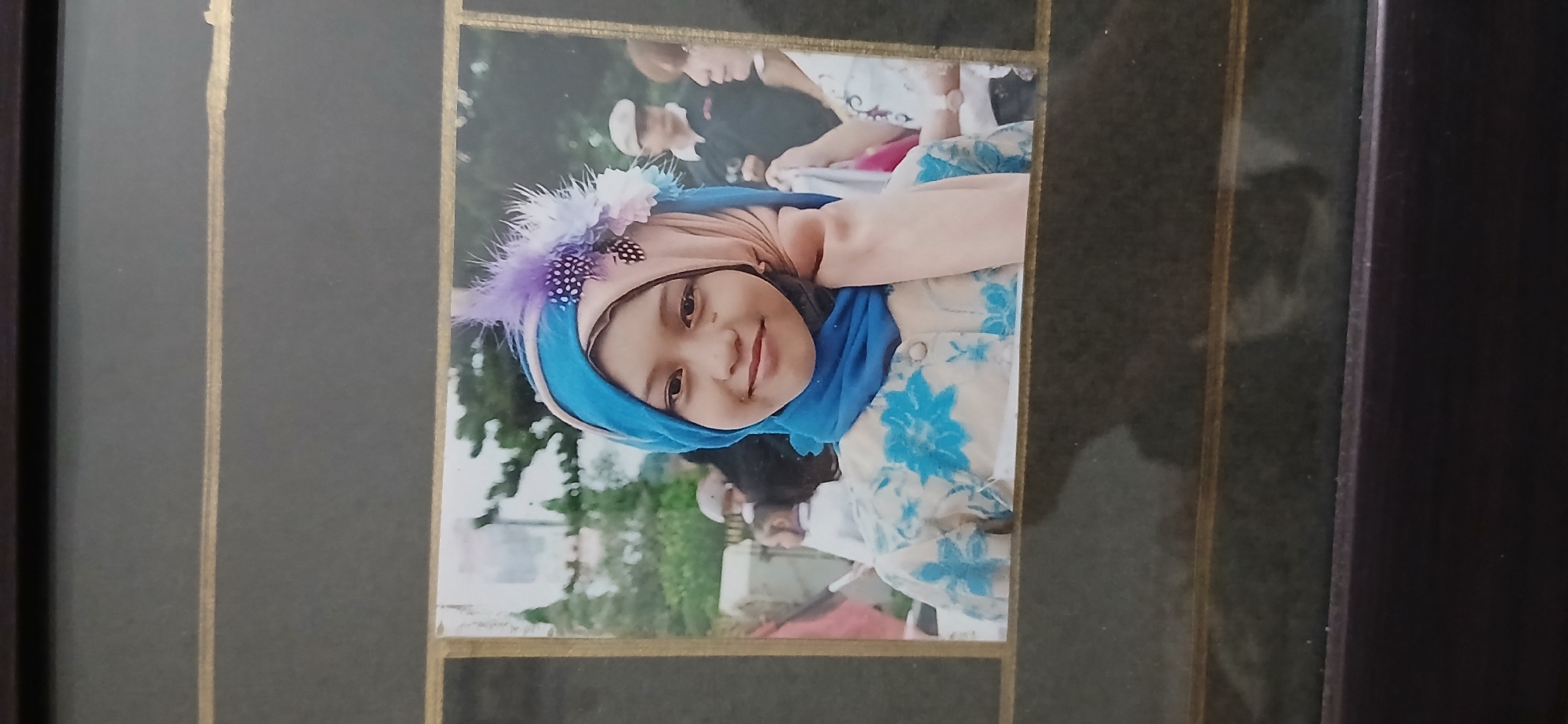 A framed photograph captures a young girl, likely Indonesian, wearing a blue and tan hijab adorned with purple and black feathers. She is dressed in a cream-colored shirt with a blue floral pattern. She smiles directly at the camera, her right hand resting near her chin. In the blurred background, other people are visible, suggesting an outdoor gathering. The location is identified as Samarinda, Indonesia.