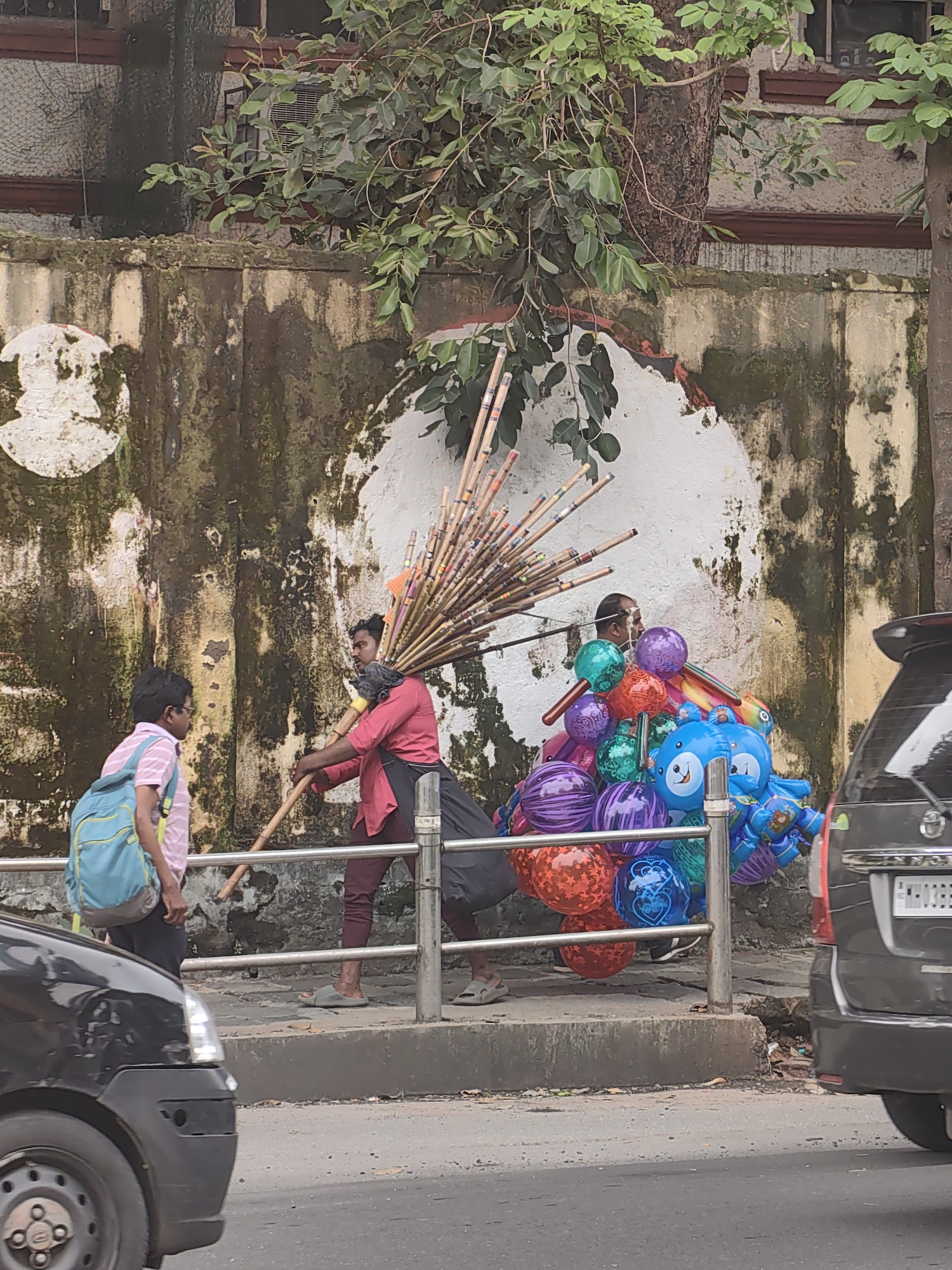 The image shows a street scene in Mumbai, India, with a man selling balloons and another person holding a collection of bamboo sticks. The man selling balloons is wearing a pink shirt and dark pants, and he is standing next to a cart filled with colorful balloons, including a large blue one shaped like a character. The other person is wearing a striped shirt and a backpack, and they are standing near the bamboo sticks. The background features a textured wall with peeling paint and some greenery, suggesting an urban environment. A black car is visible in the foreground, and a part of a grey vehicle is on the right side of the image. The ground is paved, and a metal railing separates the sidewalk from the road.